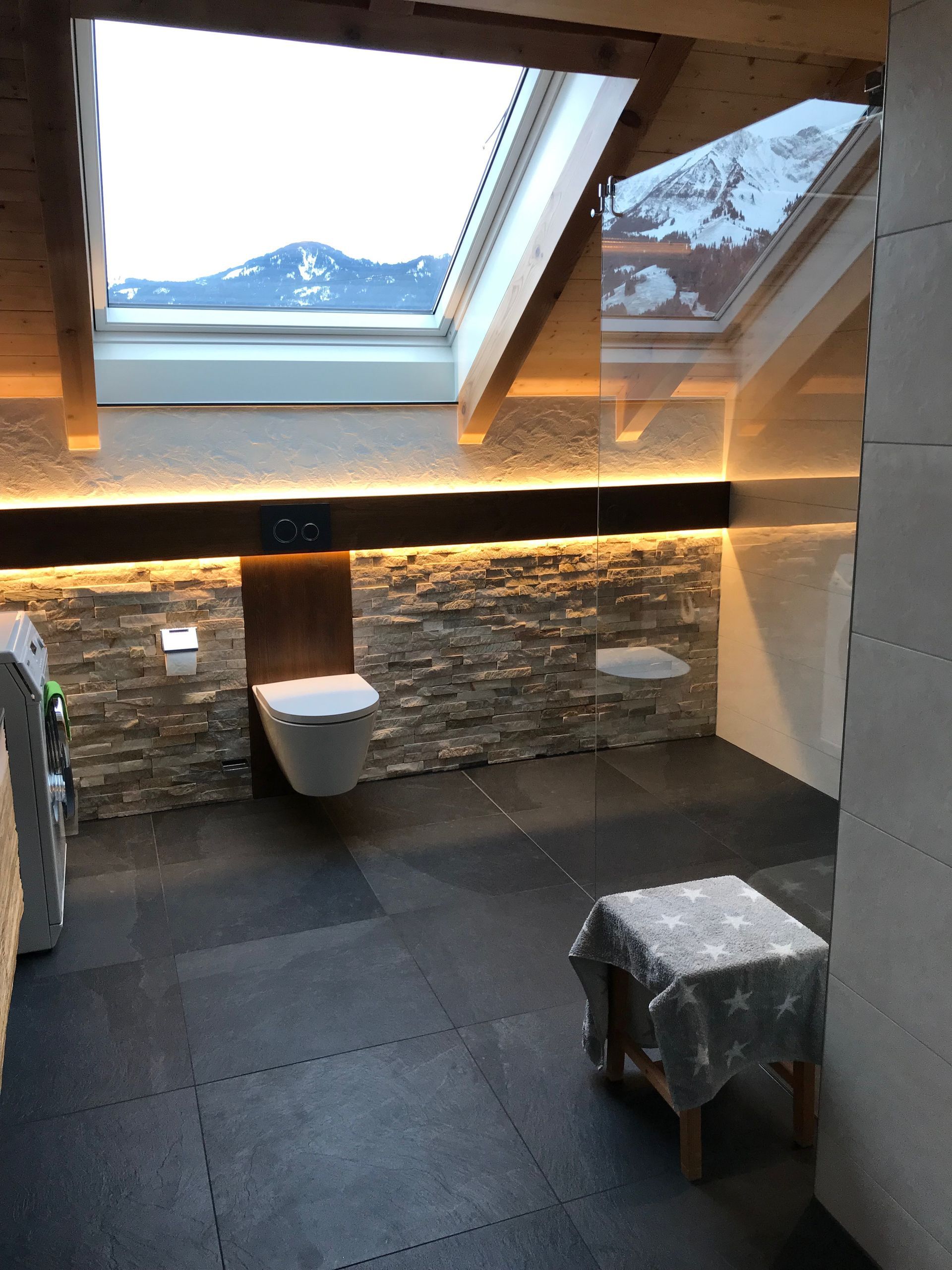 Badezimmer mit Steinwand, Oberlicht mit Bergblick, dunklem Boden, Toilette, kleinem Hocker.