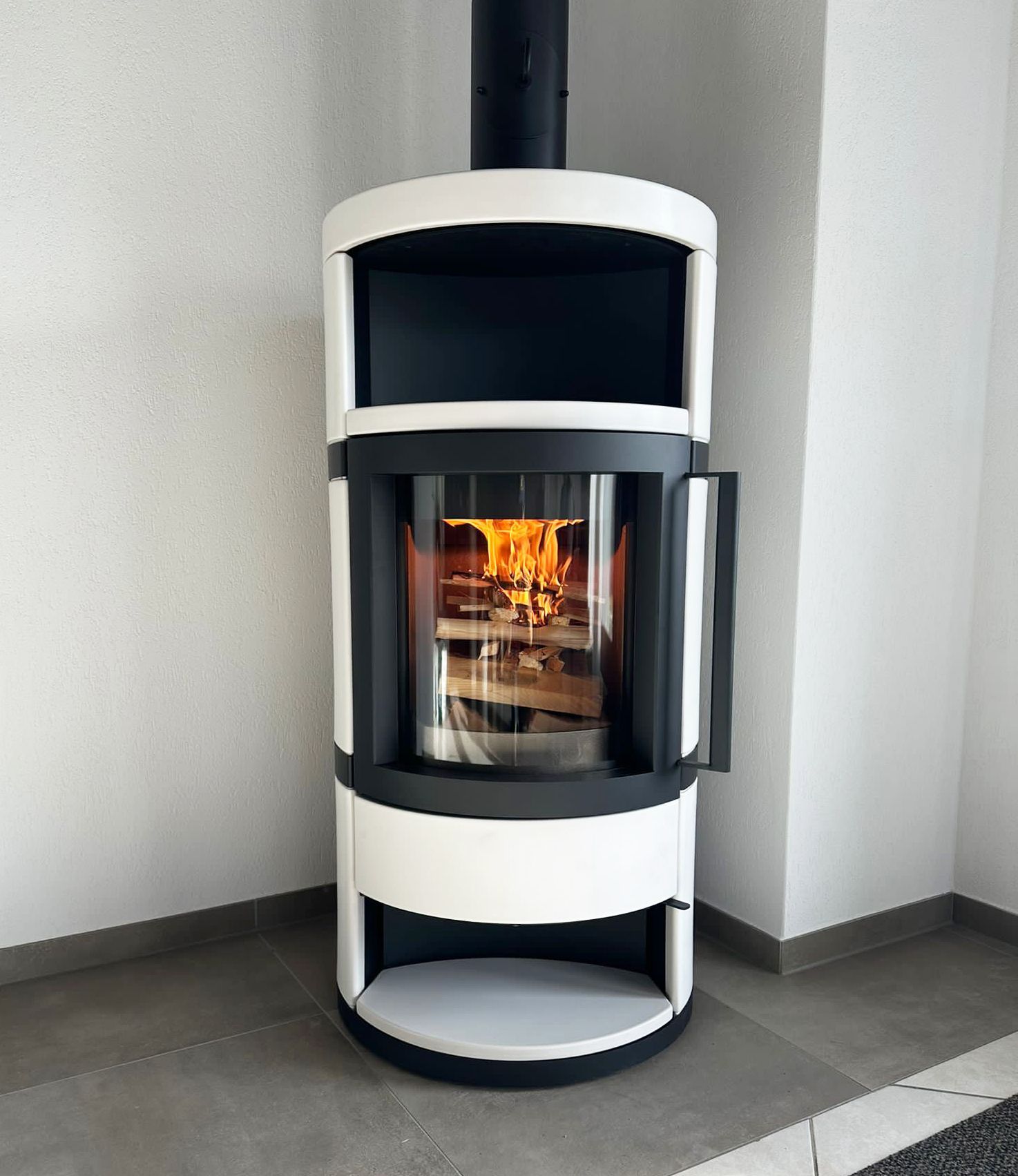 Weißer und schwarzer Holzofen mit Flammen im Inneren, Tür offen. Schwarzer Schornstein. Innenaufnahme.