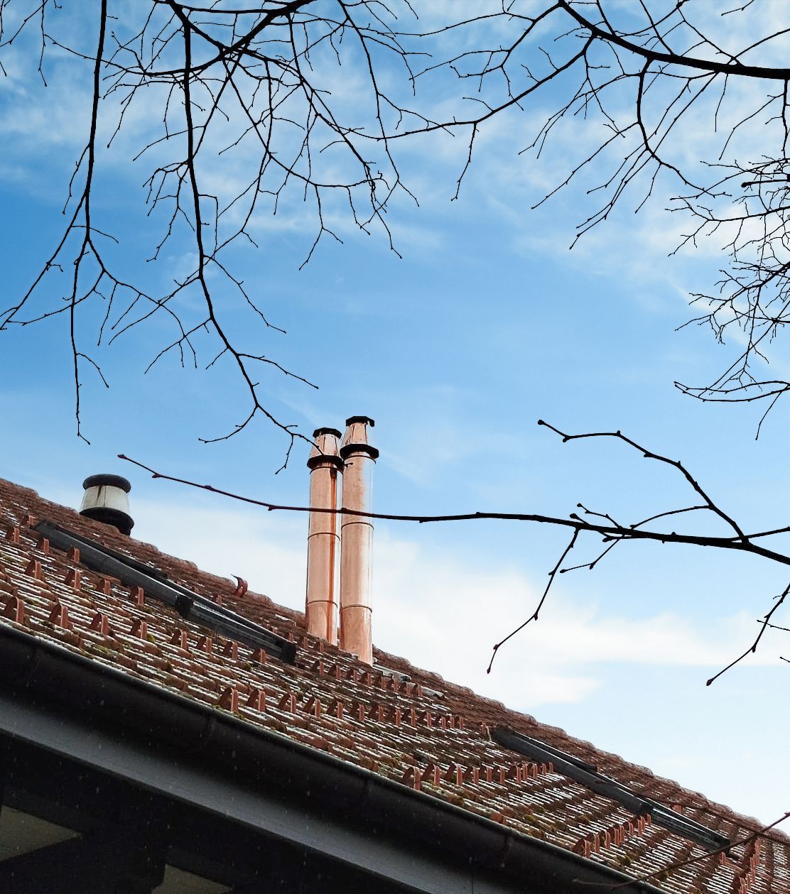 Kupferne Schornsteine ​​auf einem Ziegeldach unter blauem Himmel, eingerahmt von kahlen Baumzweigen.
