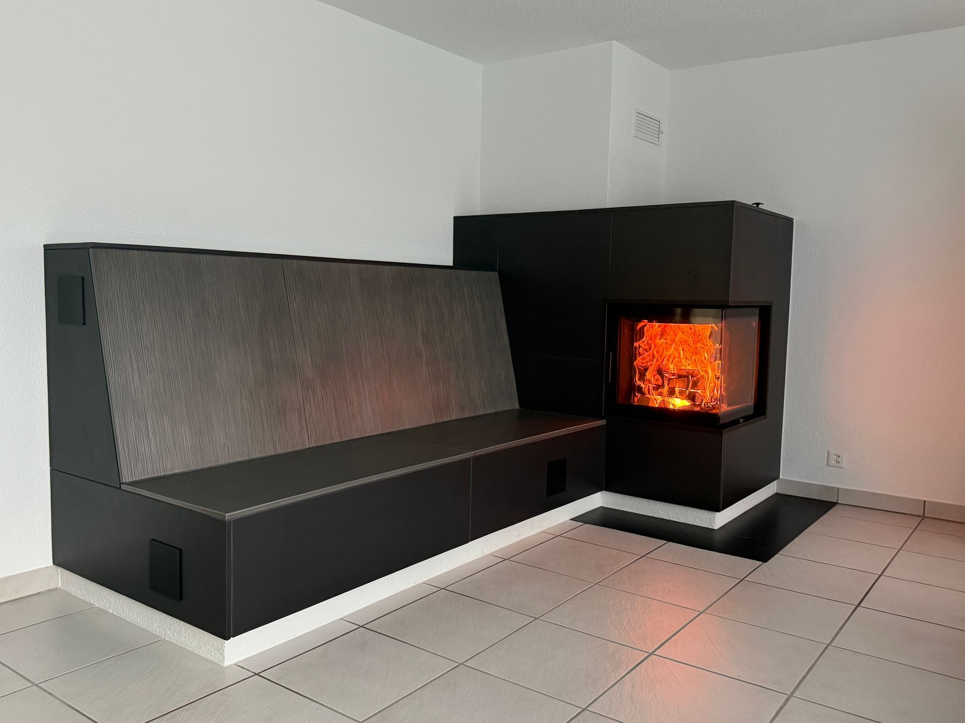 Moderne, schwarze Einbaubank mit Kamin. Im Kamin lodert ein Feuer. Weiße Fußleisten, gefliester Boden.