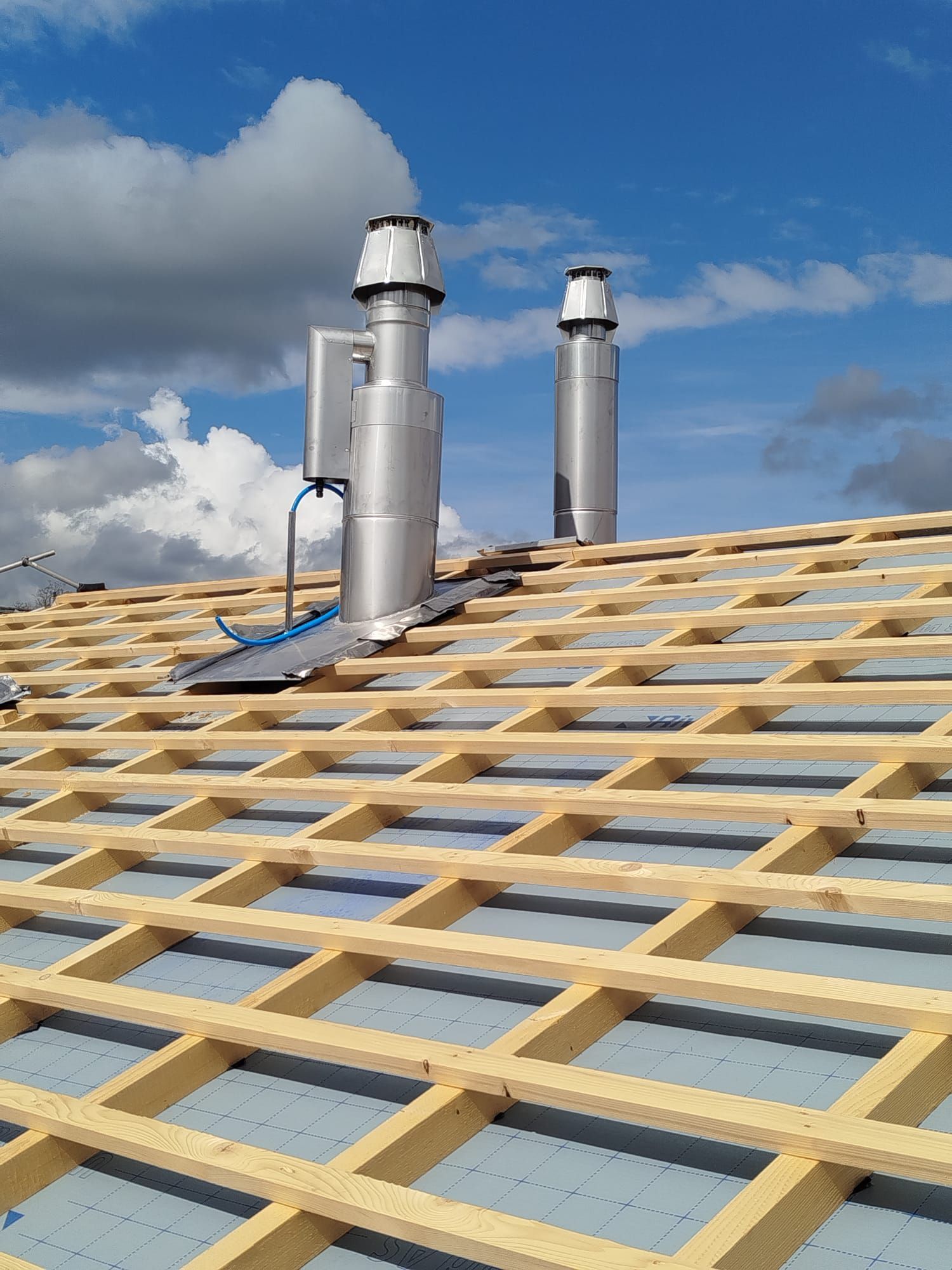 Zwei silberne Auspuffrohre auf einem Dach mit hölzernen Dachsparren und blauem Himmel mit Wolken.