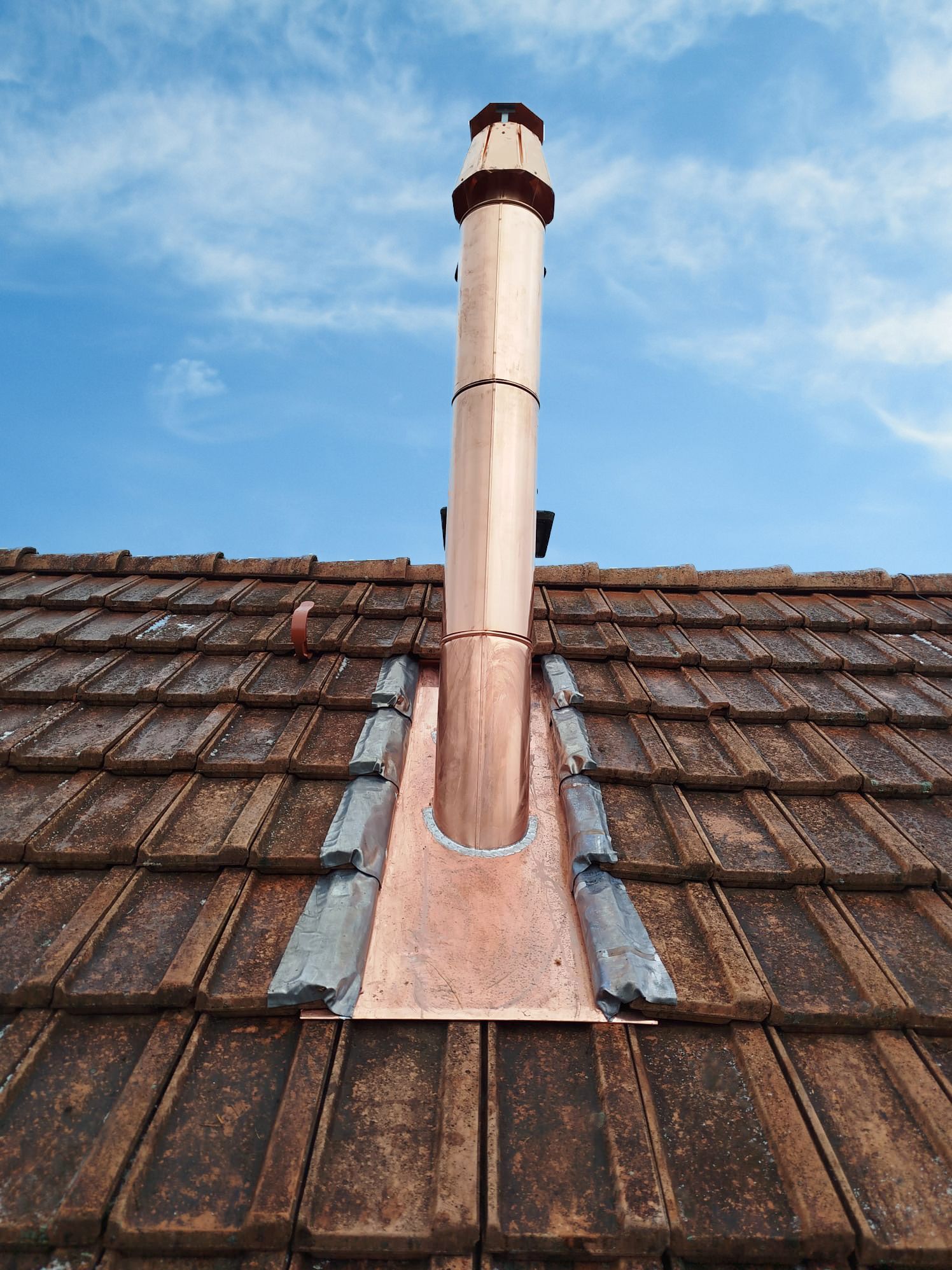 Kupferner Schornstein auf einem Ziegeldach vor blauem Himmel.
