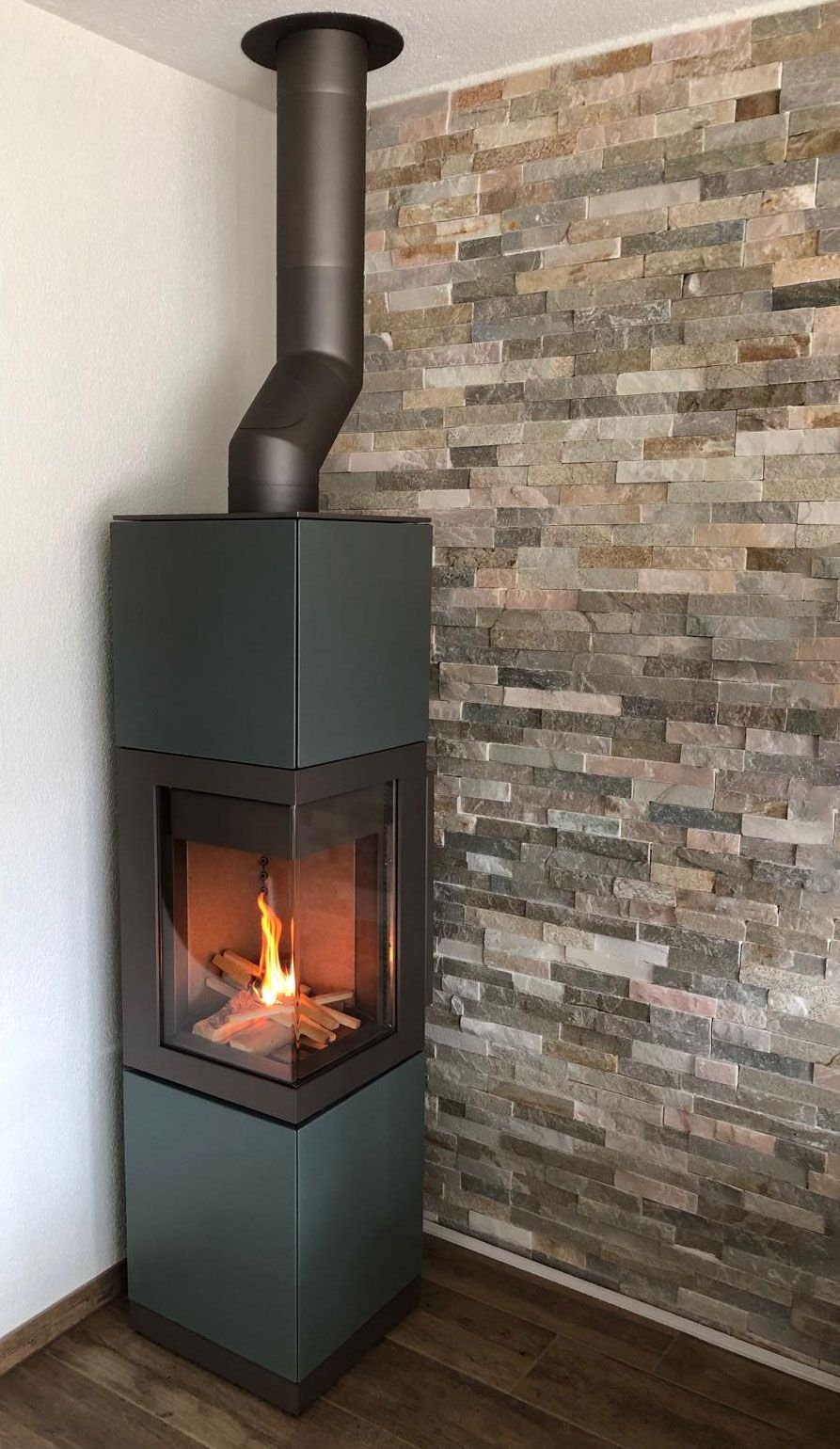 Dunkelgrüner Ofen mit brennendem Feuer, neben einer Steinmauer, brauner Schornstein.