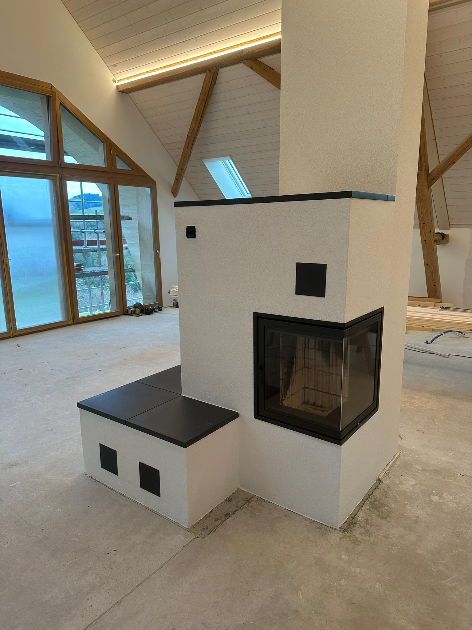 Moderner weißer Kamin mit schwarzen Akzenten vor einem Raum mit großen Fenstern und Holzbalken.