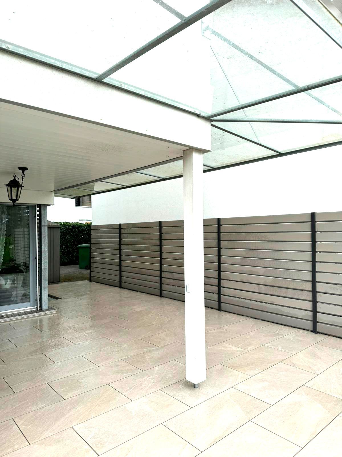 Überdachte Terrasse mit transparentem Dach, weißer Säule, grauem Lattenzaun und hellem Steinboden.