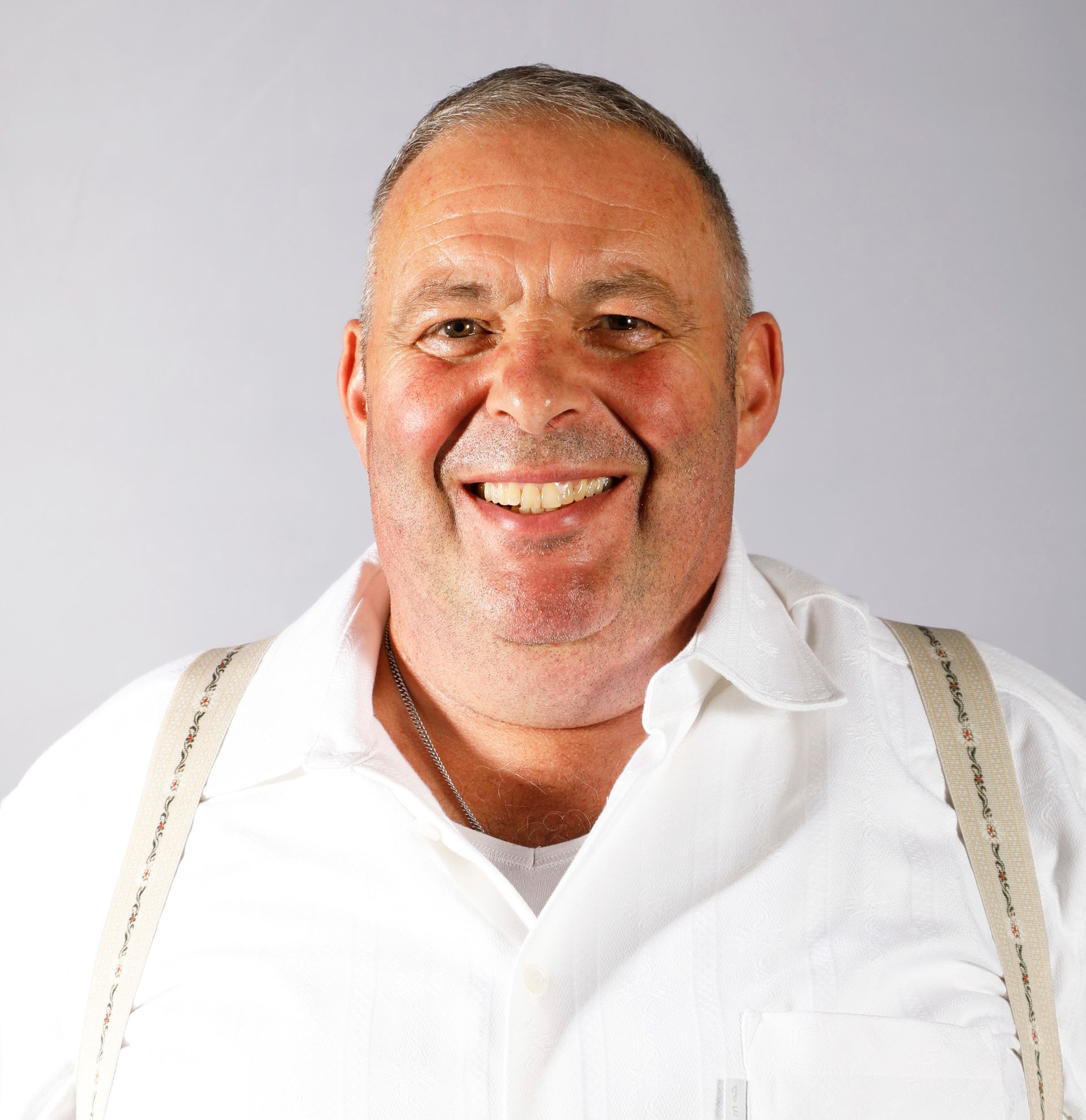 Ein Mann mit kurzen Haaren lächelt vor einem grauen Hintergrund. Er trägt ein weißes Hemd und Hosenträger.