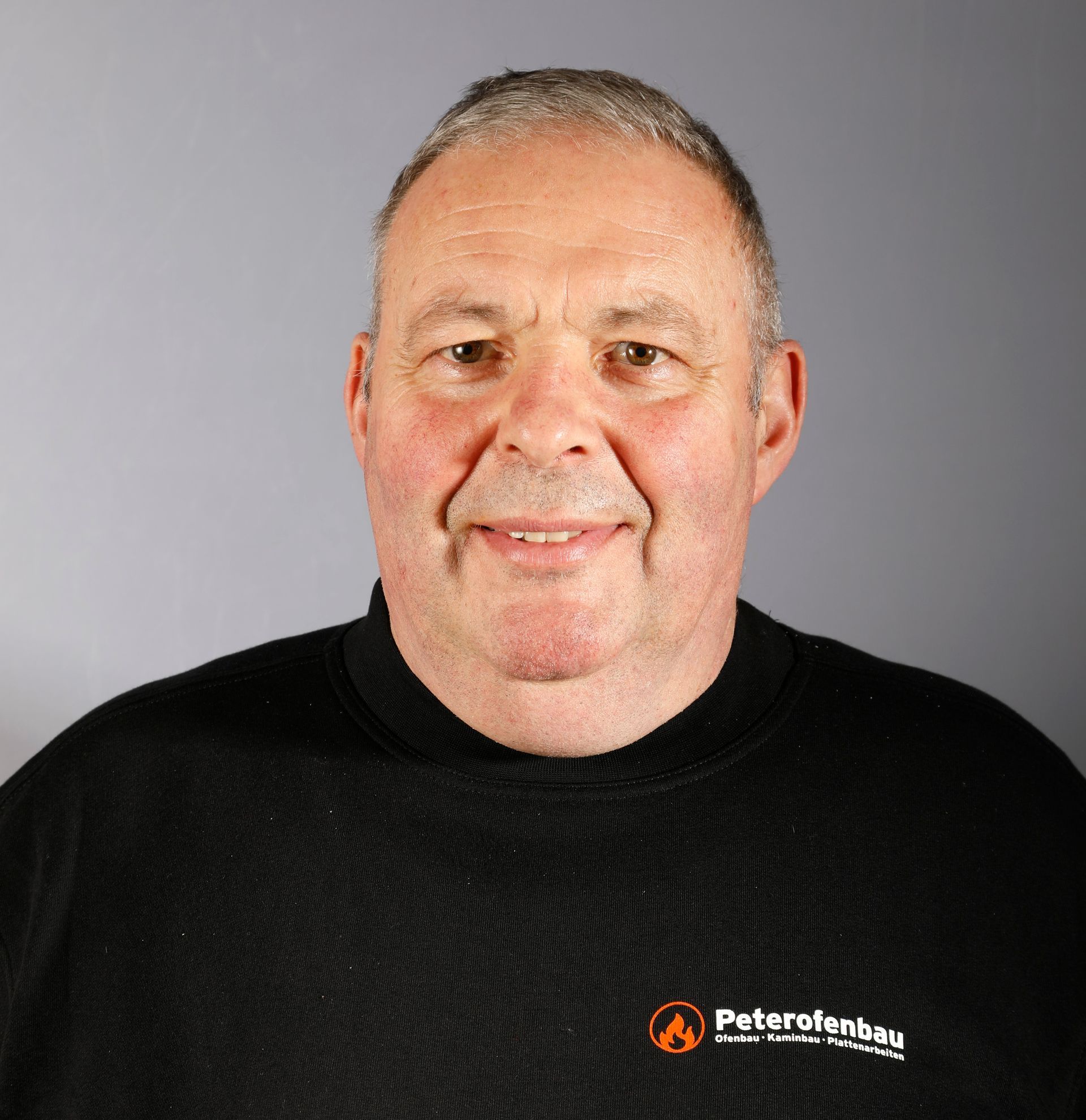 Ein Mann mit kurzen Haaren lächelt vor einem grauen Hintergrund. Er trägt ein weißes Hemd und Hosenträger.