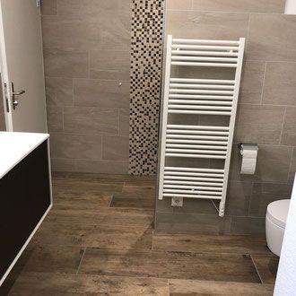 Badezimmer mit Fliesenboden in Holzoptik, grauen Fliesenwänden, einem weißen Handtuchwärmer und einer Toilette.