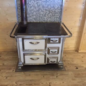 Weißer und silberner Holzofen mit Schubladen, Kochfeld und Schornstein vor Mosaikfliesen und Holzvertäfelung.