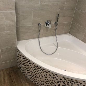Eckbadewanne mit Mosaikfliesenverkleidung, Chromarmaturen und neutralfarbigen Fliesenwänden.