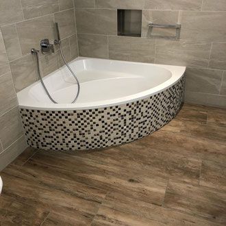 Eckbadewanne mit Mosaikfliesensockel, Chromarmaturen und Fliesenboden in Holzoptik.