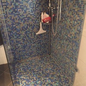 Duschkabine mit blauen Mosaikfliesen an Wänden und Boden, Duschkopf und kleinem Regal.