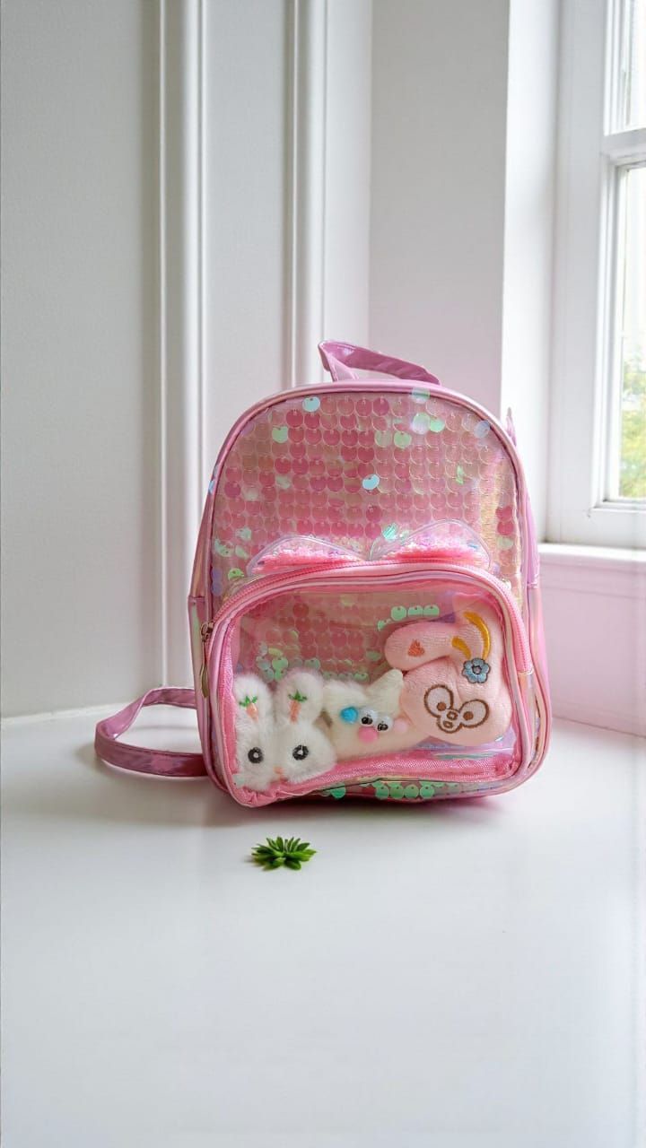 Mochila rosa con lentejuelas y un bolsillo frontal transparente donde se guardan peluches, junto a una ventana.