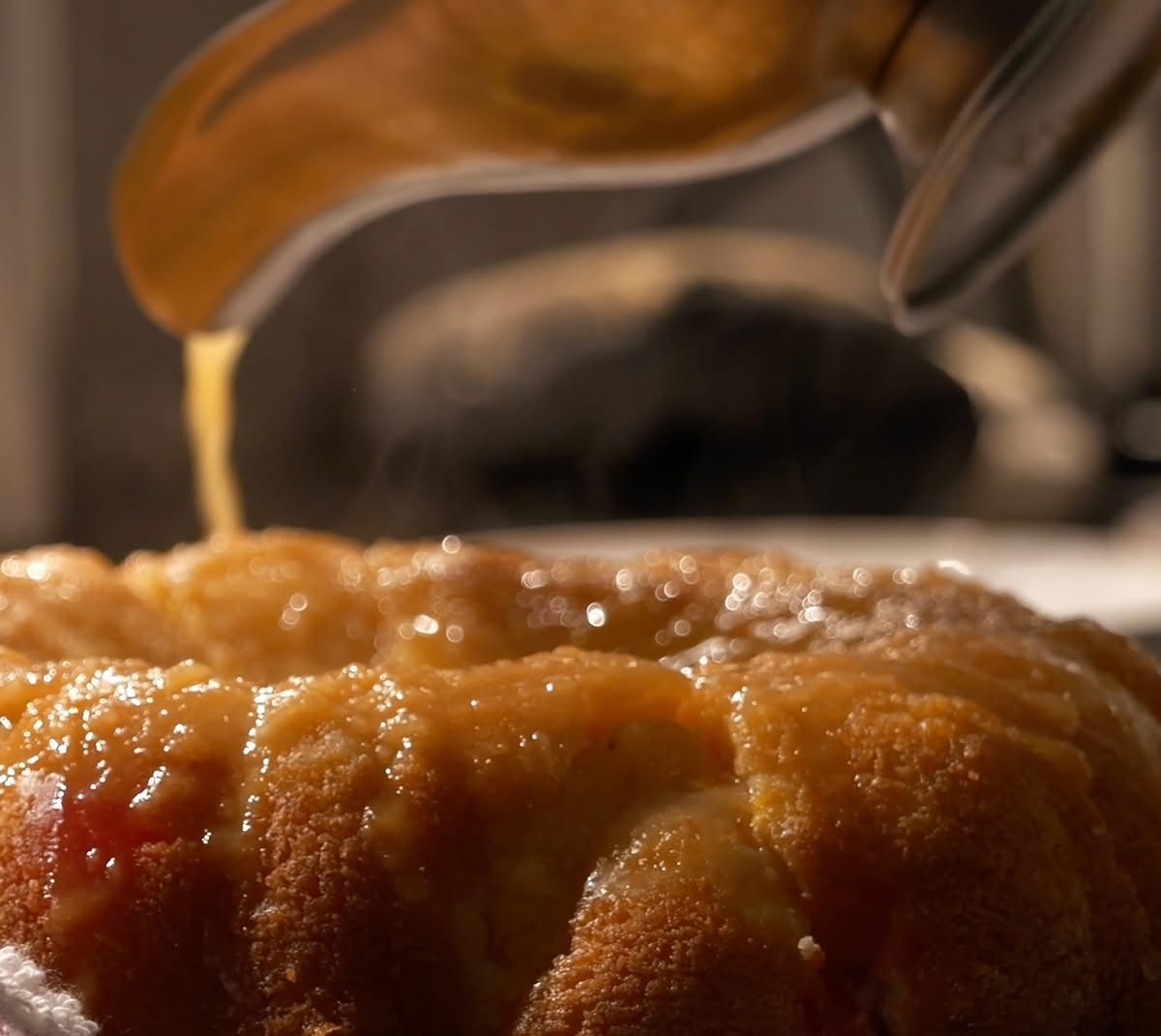 Pineapple Rum Cake being drizzled with a golden syrup. Close-up with focus on cake, steam rising, and pouring spout.