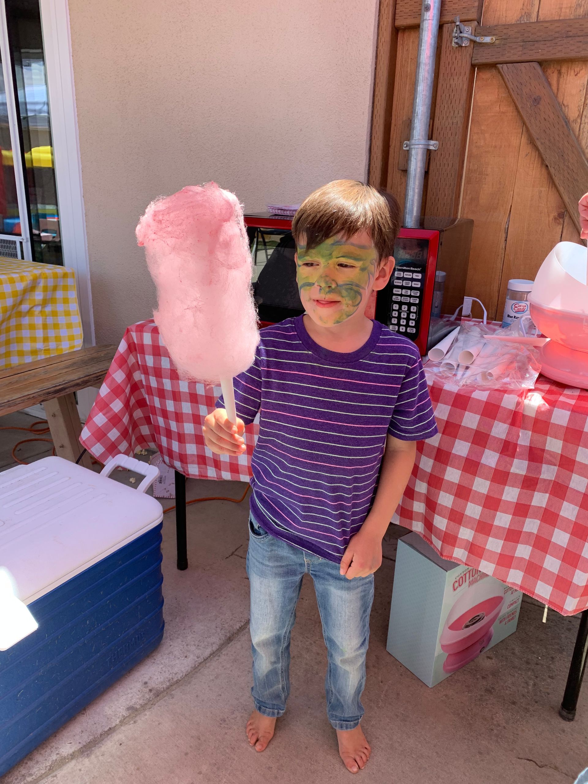 A young boy with paint on his face is eating cotton candy.