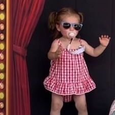 A little girl wearing sunglasses and a pacifier in her mouth is standing in front of a red curtain.