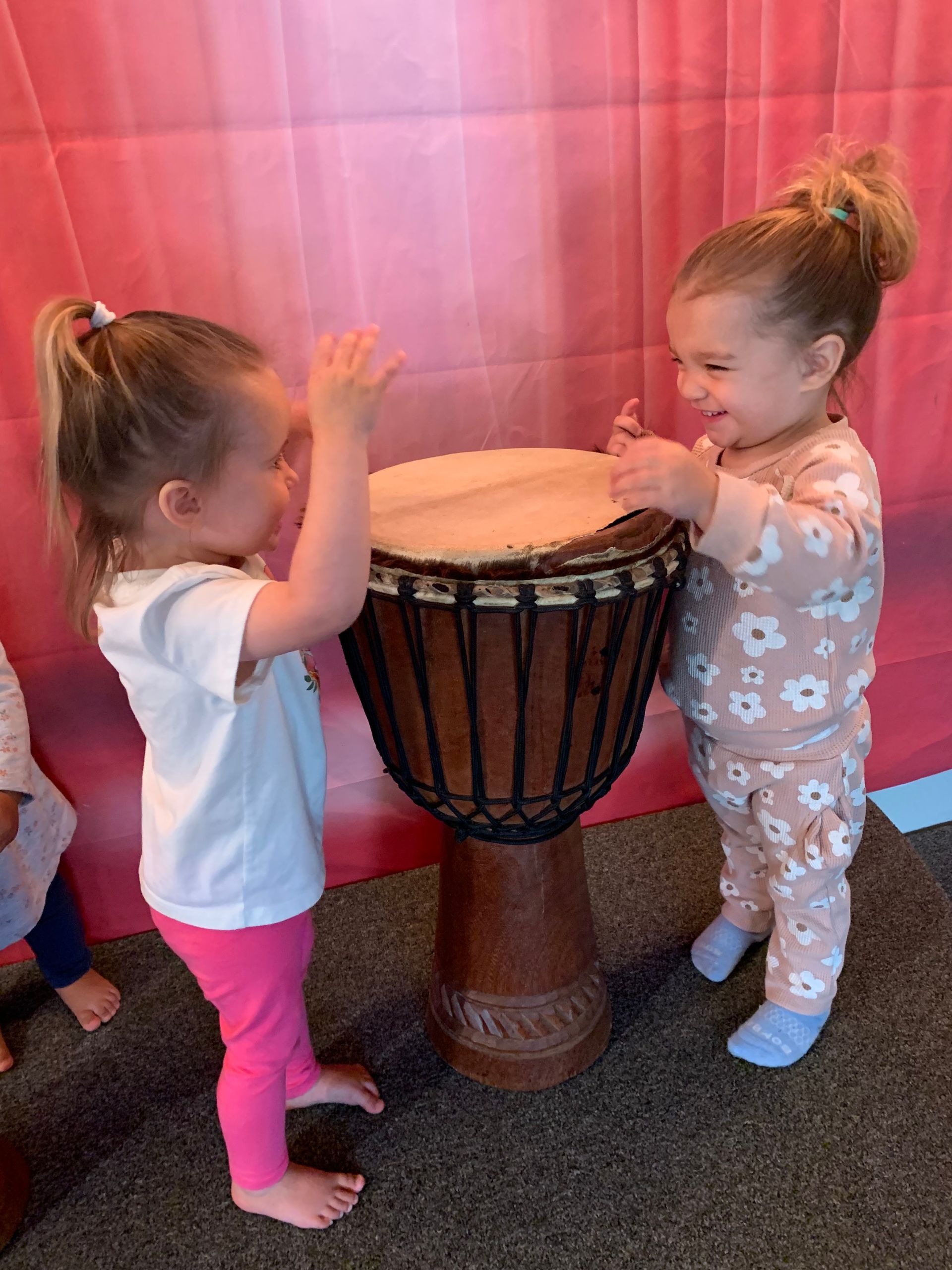 Two little girls are playing a djembe drum in a room.