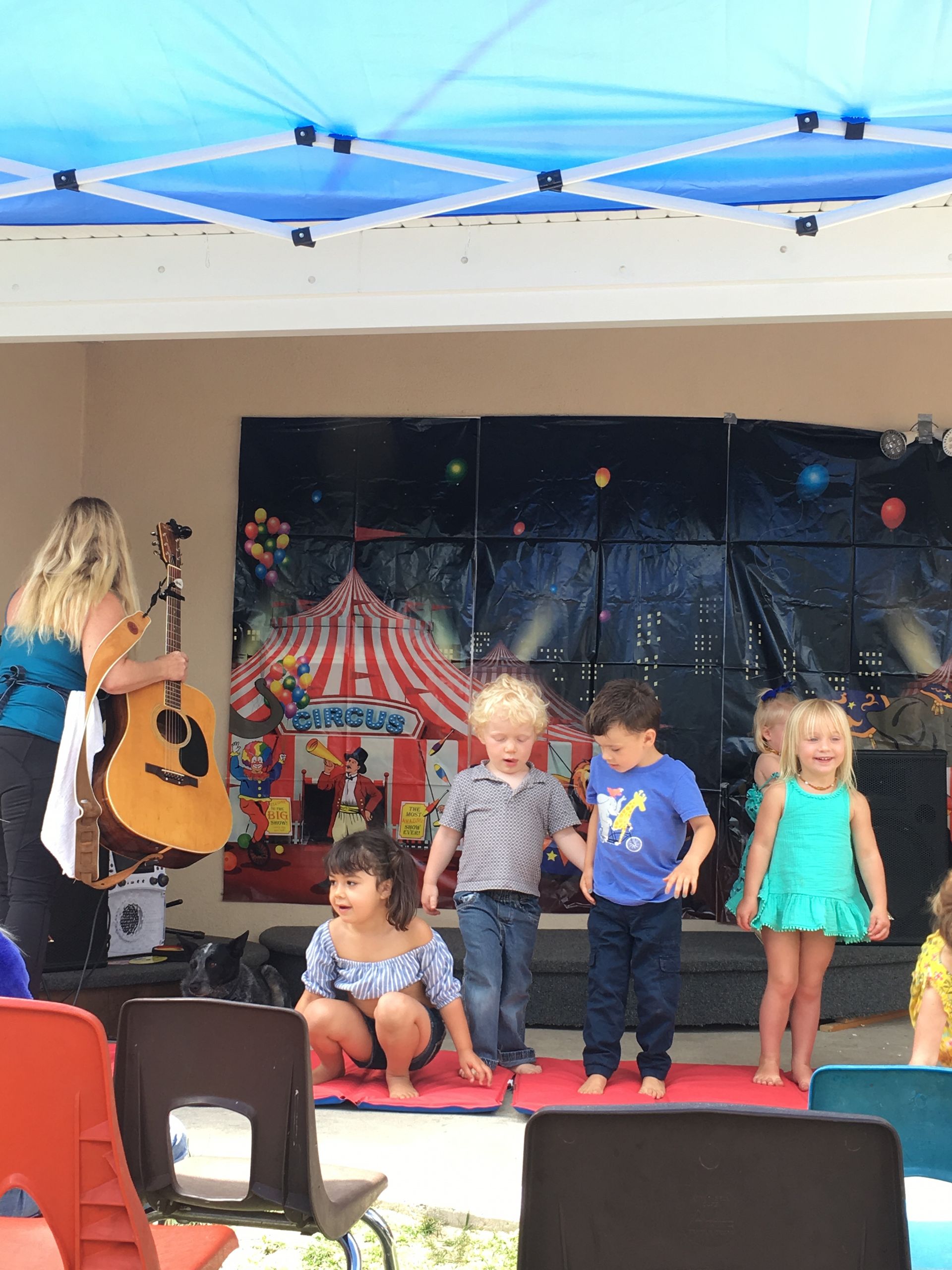 A group of children are standing on a stage while a woman plays a guitar.