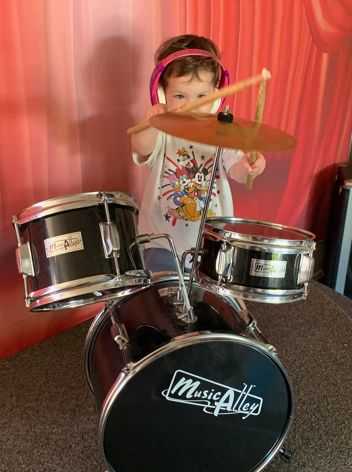 A little girl is playing drums in front of a red curtain.