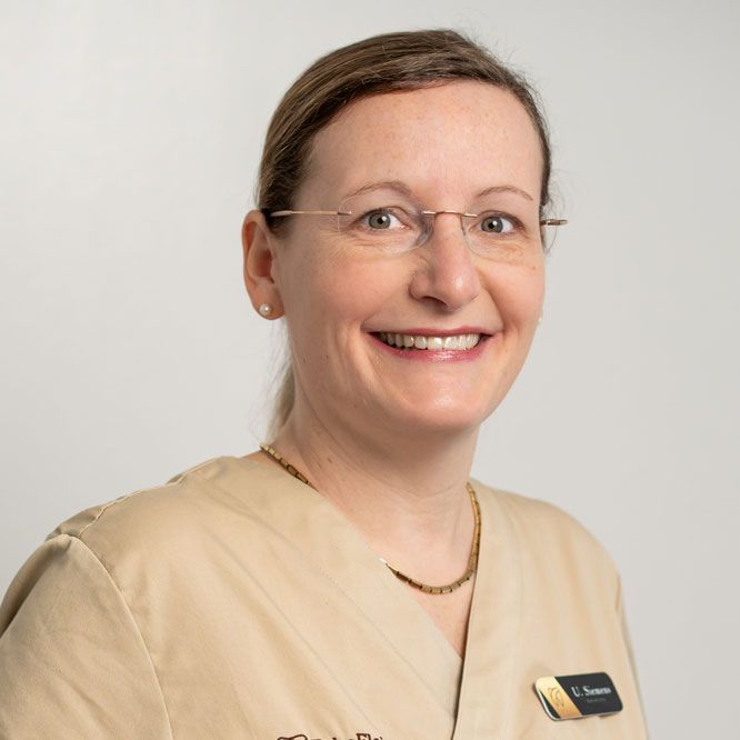Zahnärztin und Implantologie-Expertin Ute Siemens in beiger medizinischer Uniform und Brille mit Namensschild vor einem schlichten hellgrauen Hintergrund.