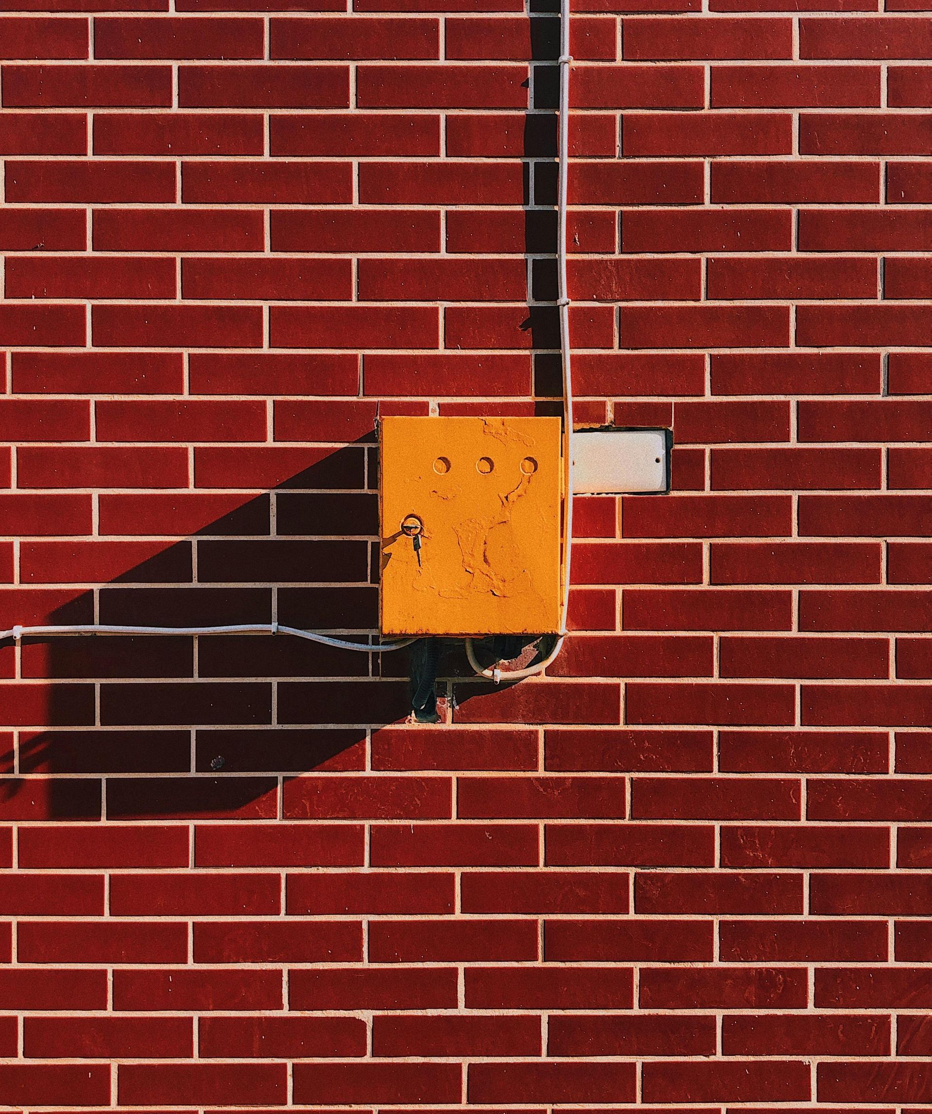 Yellow box on a red brick wall, casting a long shadow from a sunny angle.