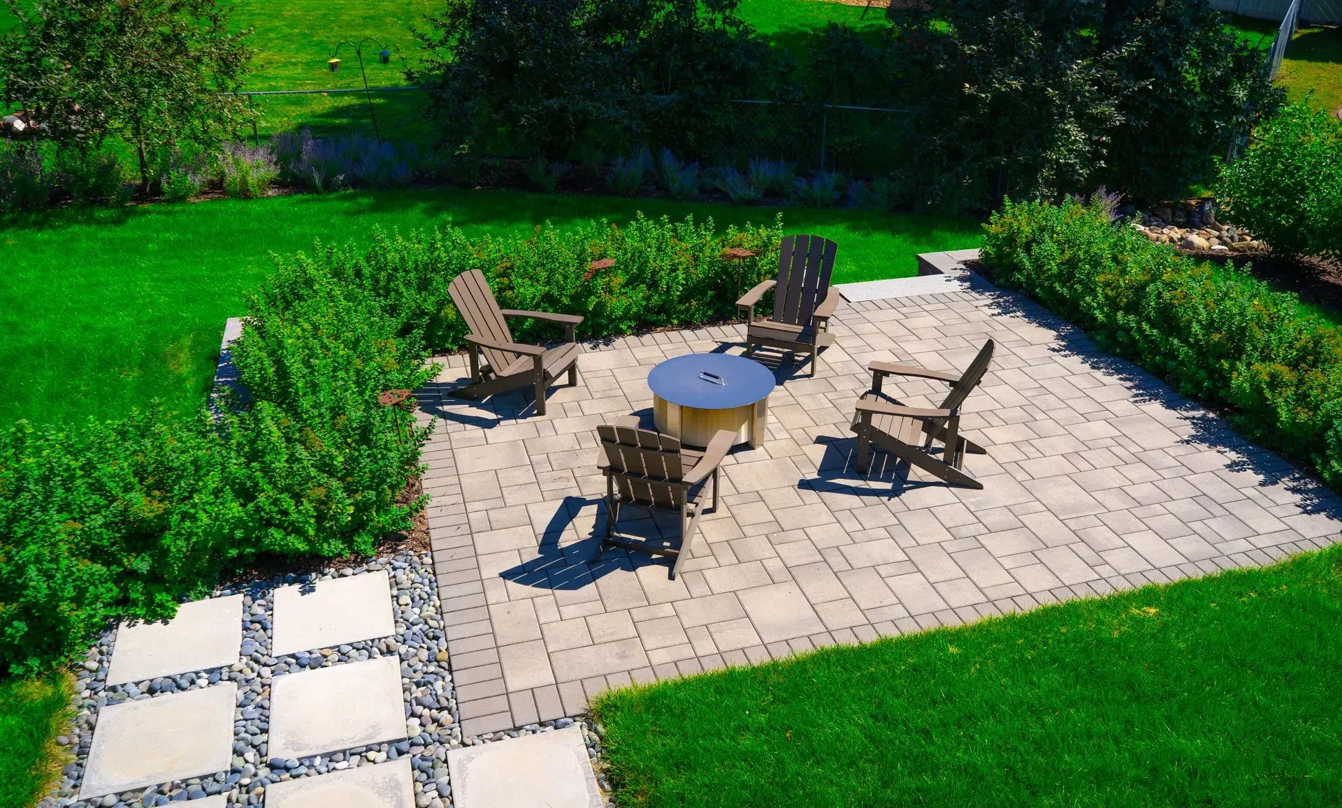 A paved patio with chairs and a fire pit sits surrounded by green lawn and landscaped shrubs.