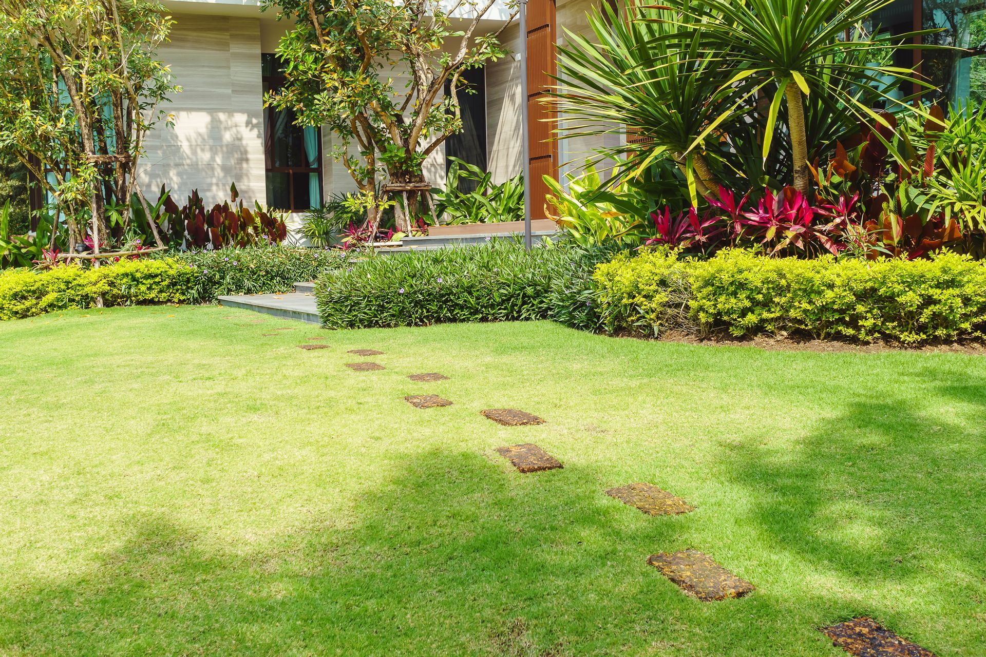 A stone stepping stone path leads across a manicured green lawn toward a modern house with lush tropical landscaping. A stone stepping stone path leads across a manicured green lawn toward a modern house with lush tropical landscaping.