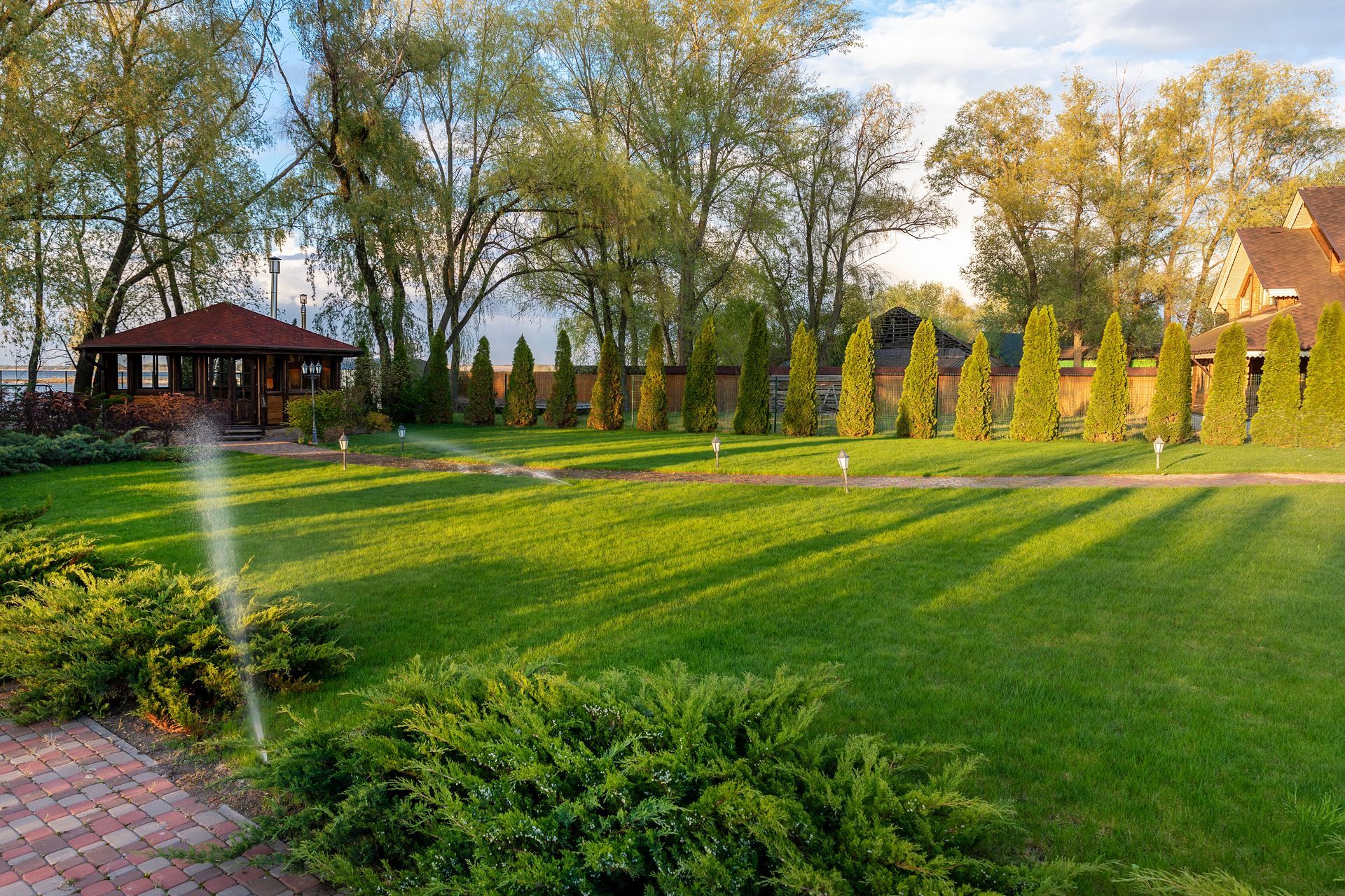A lush green lawn with a wooden gazebo and a line of evergreen trees under a soft, golden sunset light.