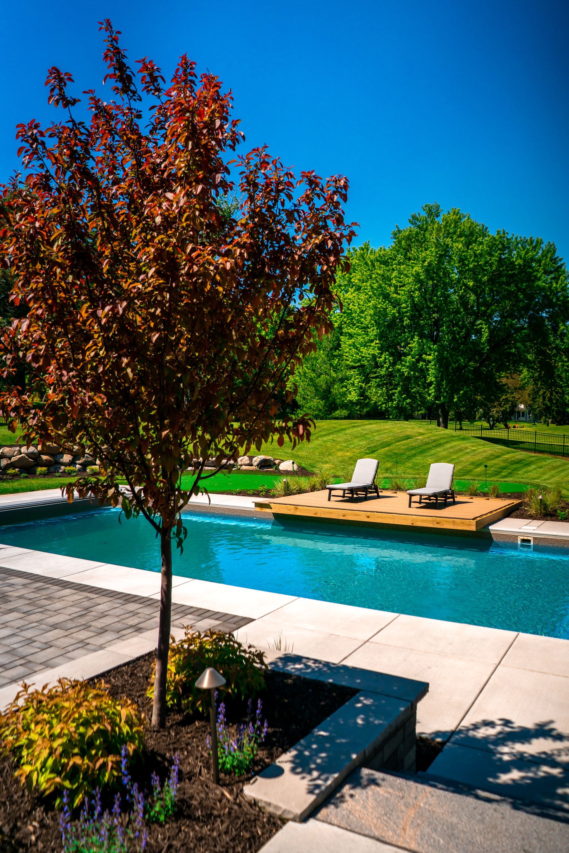 A swimming pool with a tree in the foreground and chairs in the background.