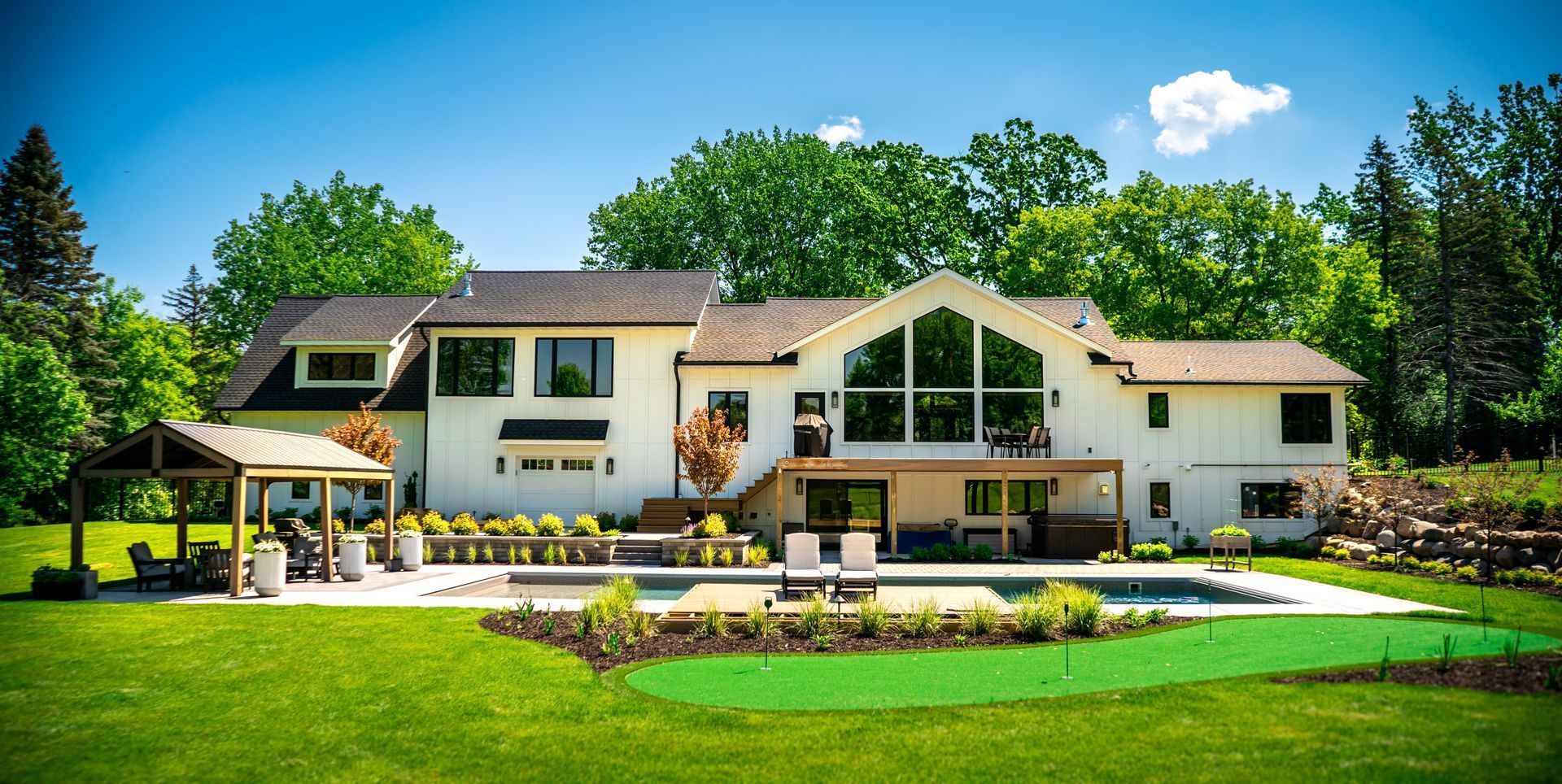 Backyard of a white house with a swimming pool, lawn, pergola, and lush green trees under a blue sky. Backyard of a white house with a swimming pool, lawn, pergola, and lush green trees under a blue sky.