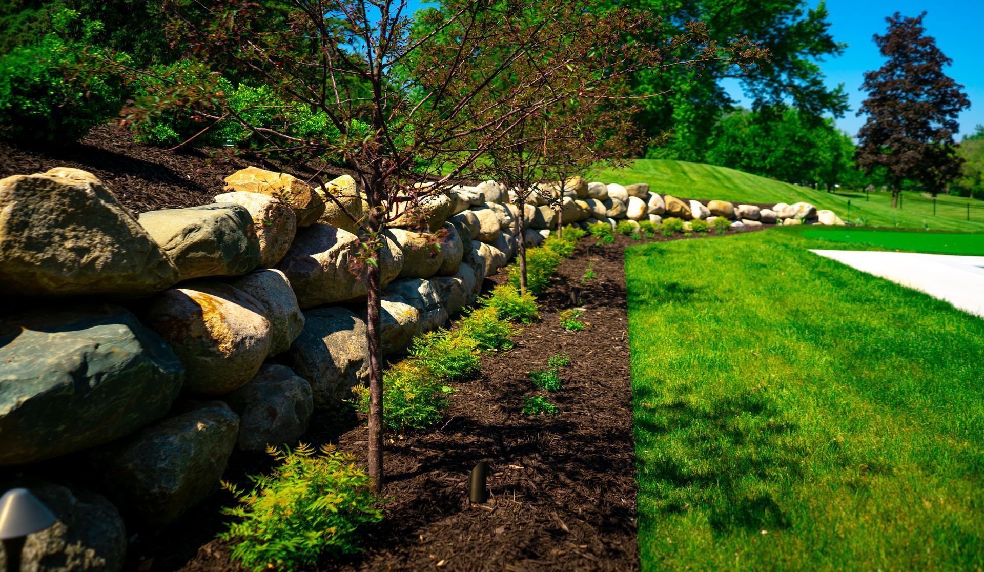 Stone retaining wall separates lawn from garden with small trees and bushes. Stone retaining wall separates lawn from garden with small trees and bushes.