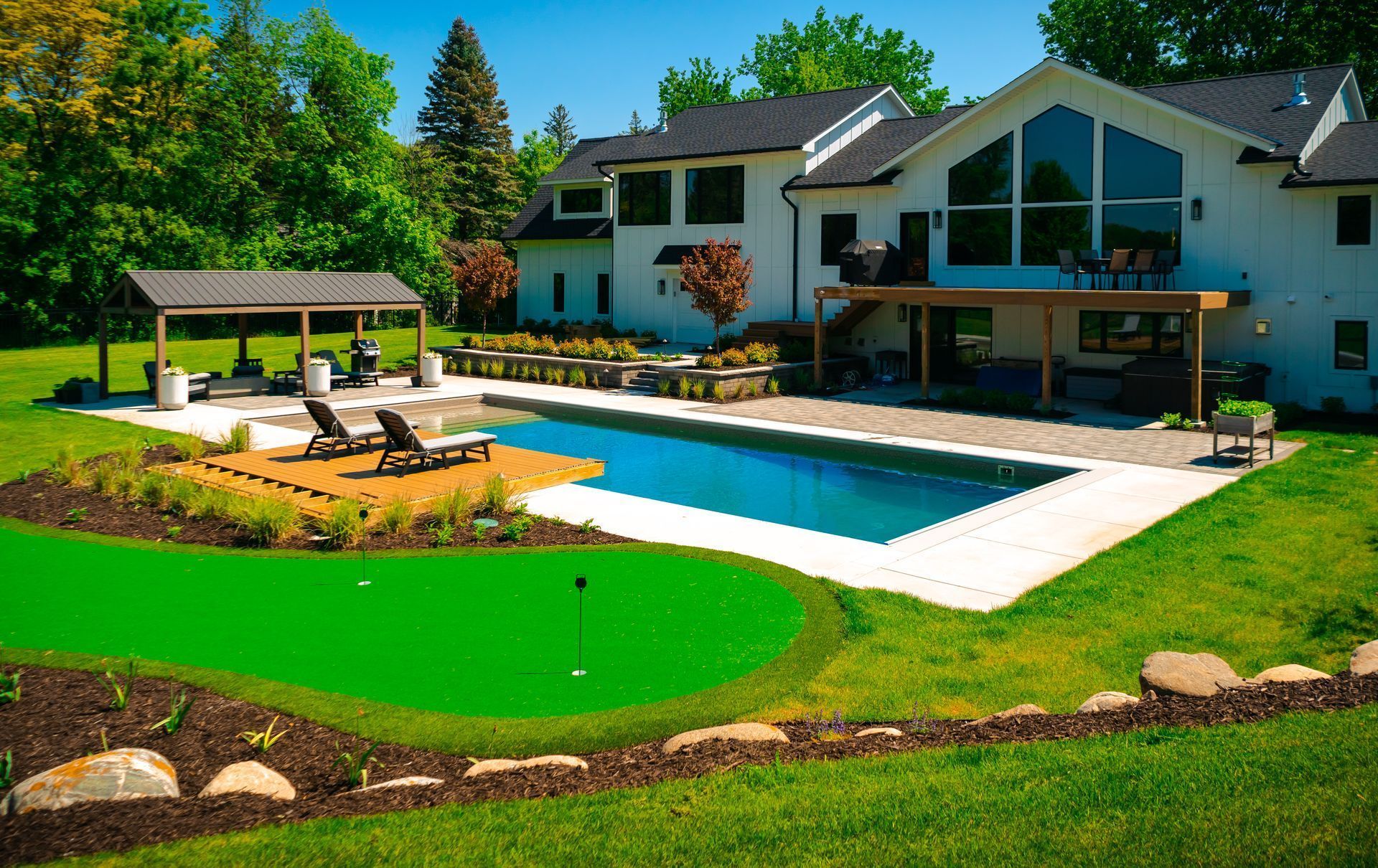 Backyard with a pool, putting green, gazebo, and white house on a sunny day. Backyard with a pool, putting green, gazebo, and white house on a sunny day.