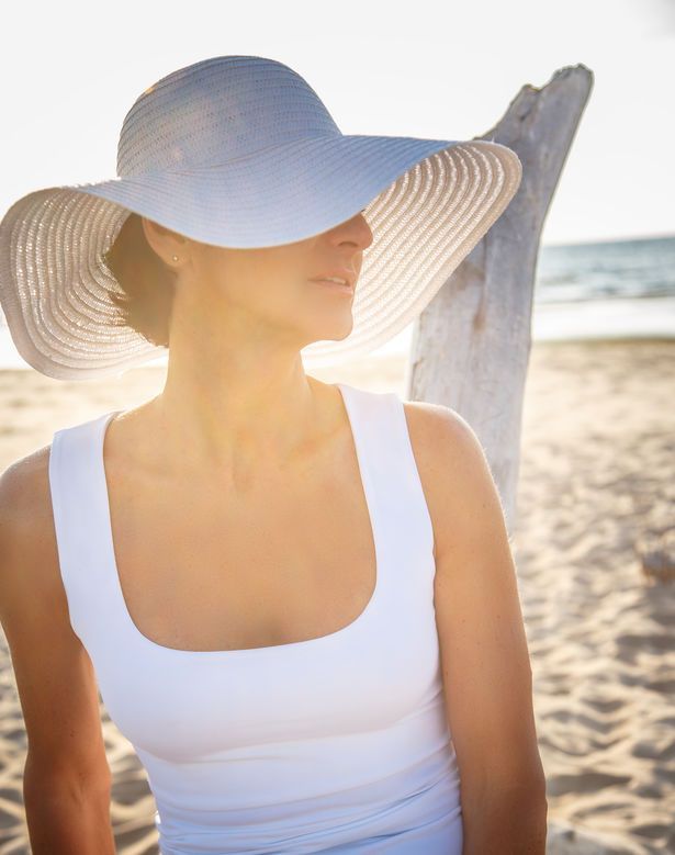 A woman wearing a white hat and a white tank top is standing on the beach.