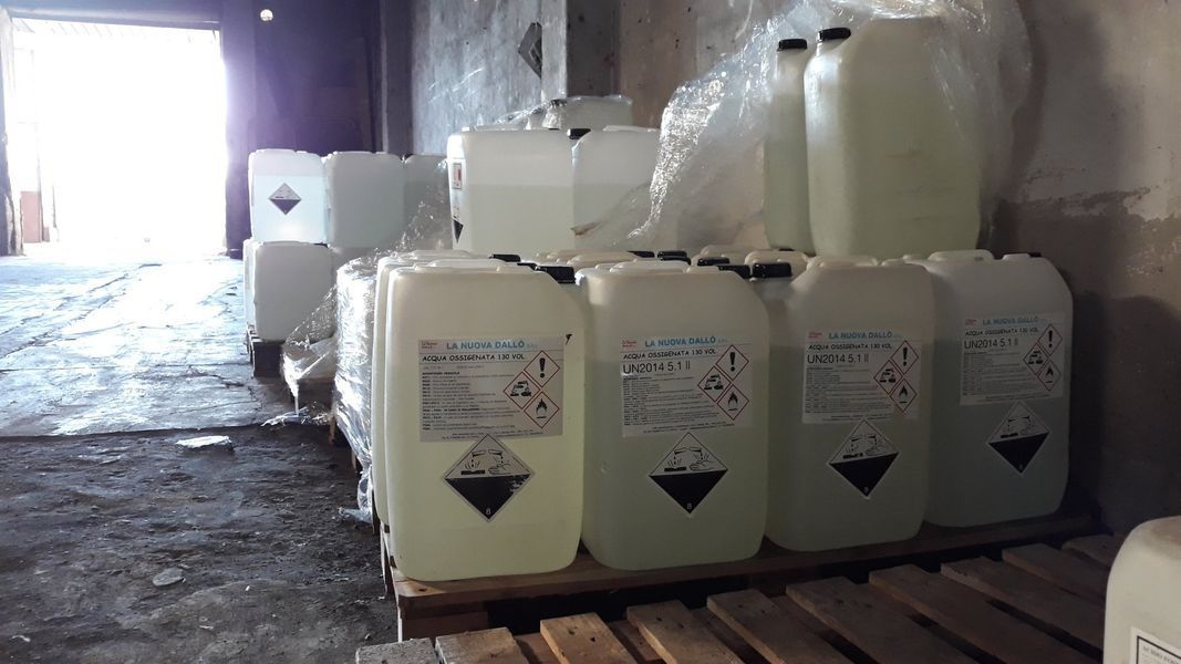 Contenitori di sostanze chimiche impilati su un pallet in un magazzino; alcuni presentano simboli di pericolo.