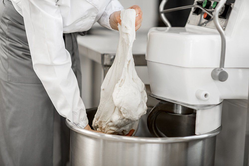 Persona con grembiule che tiene in mano l'impasto da una grande ciotola vicino a un'impastatrice commerciale.