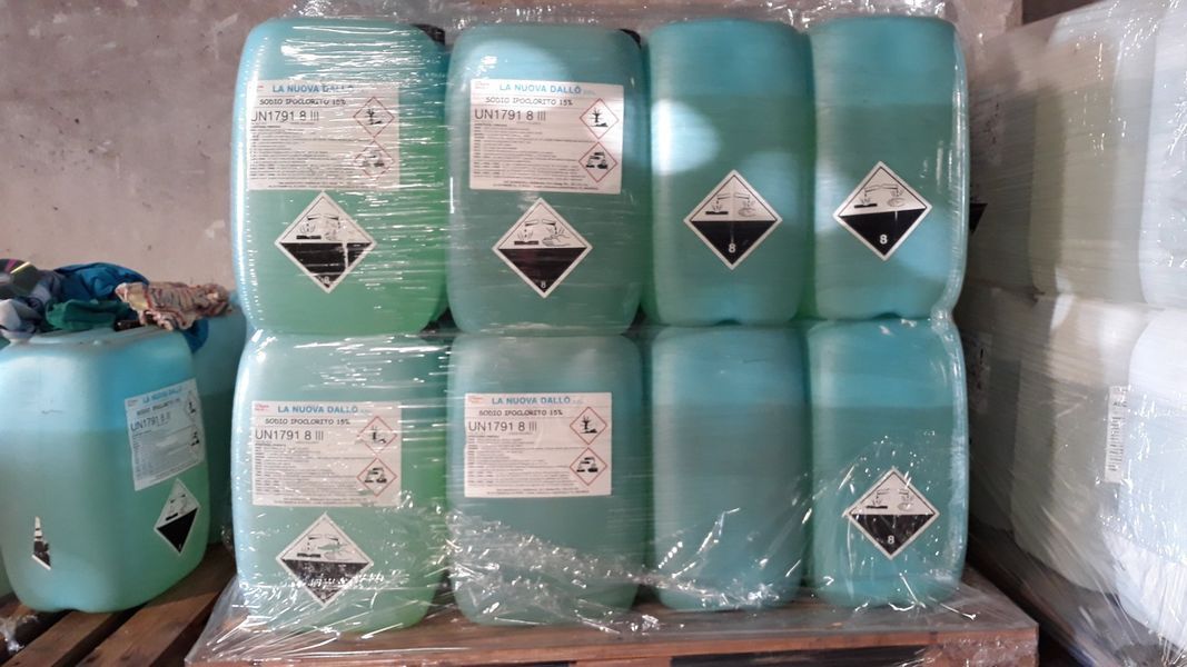 Otto contenitori di plastica blu impilati, con etichette di pericolo, avvolti nella plastica e disposti su un pallet.