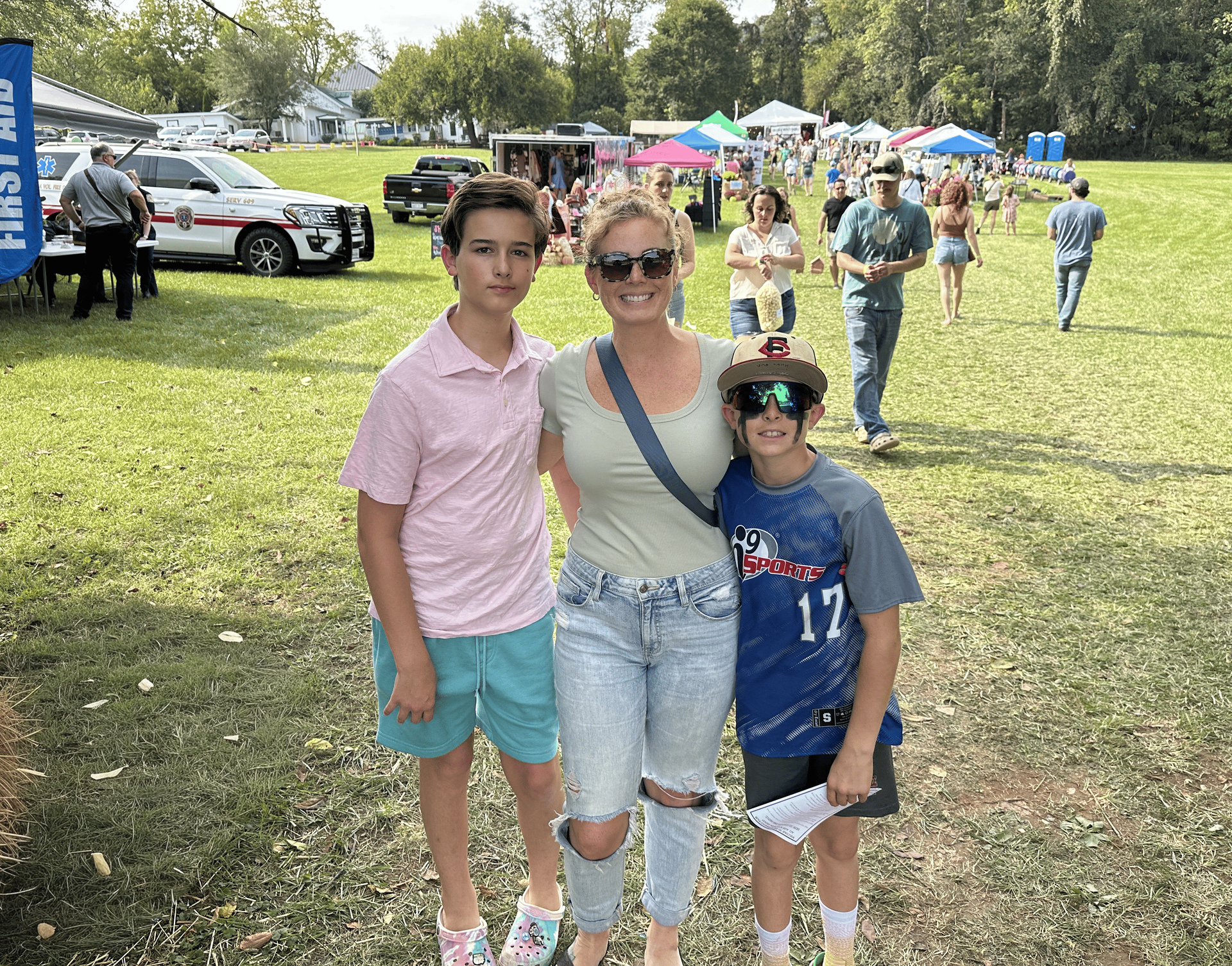 A woman and two boys smiling in a field; an outdoor event is in the background.