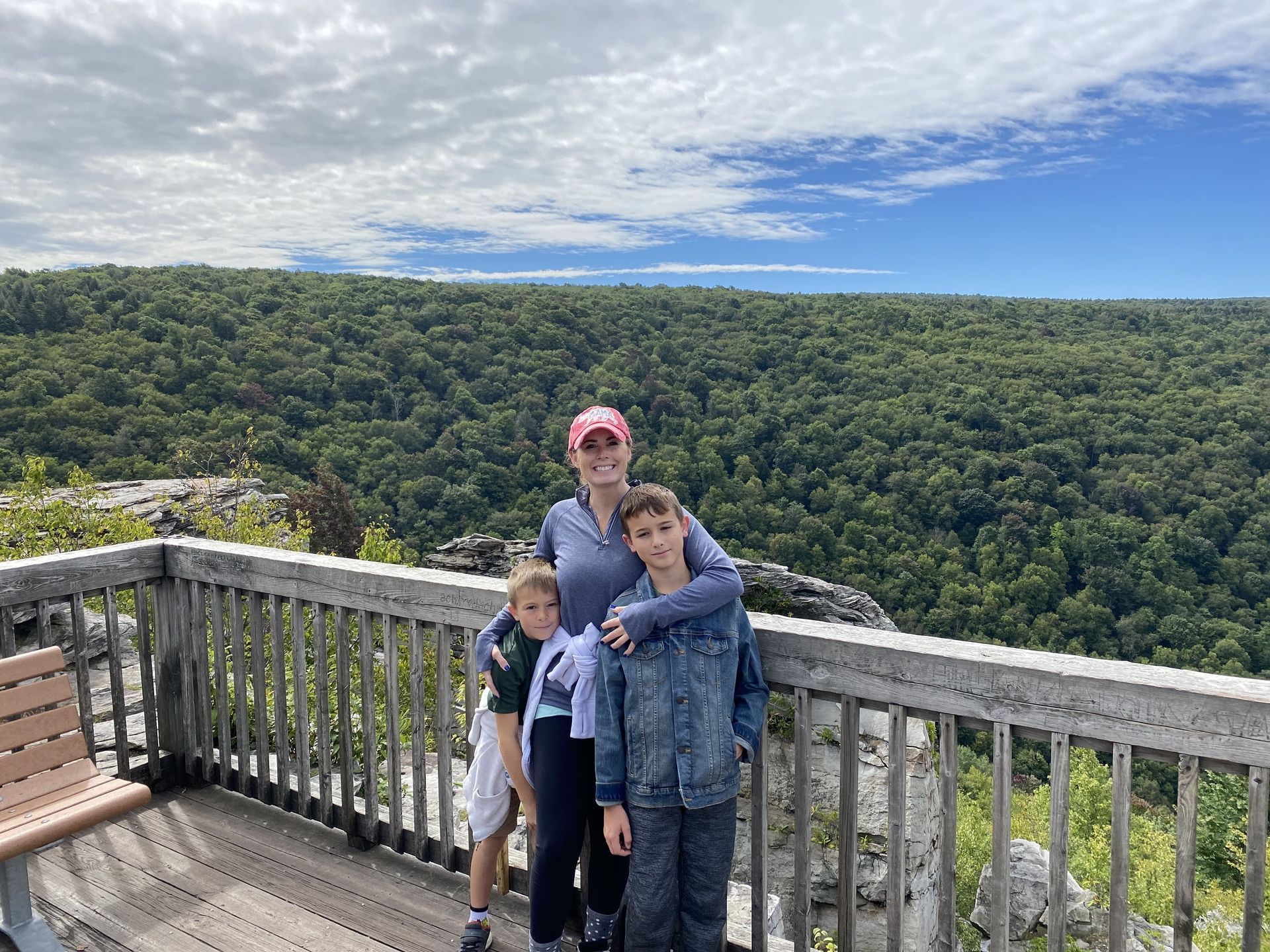 Woman and two boys on a wooden overlook, hugging, overlooking lush green forest under a blue sky with clouds.