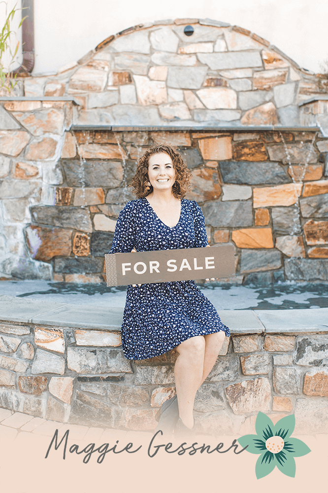 A woman is sitting on a stone wall holding a for sale sign.