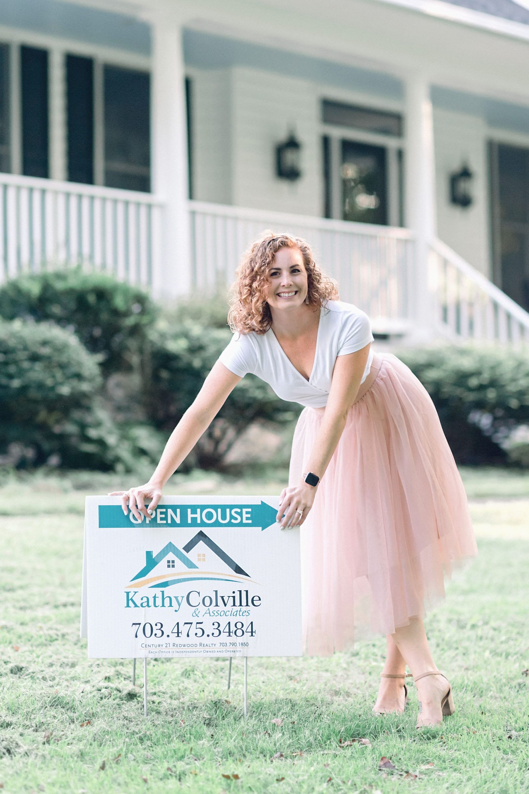 A woman in a pink skirt is holding an open house sign in front of a house.