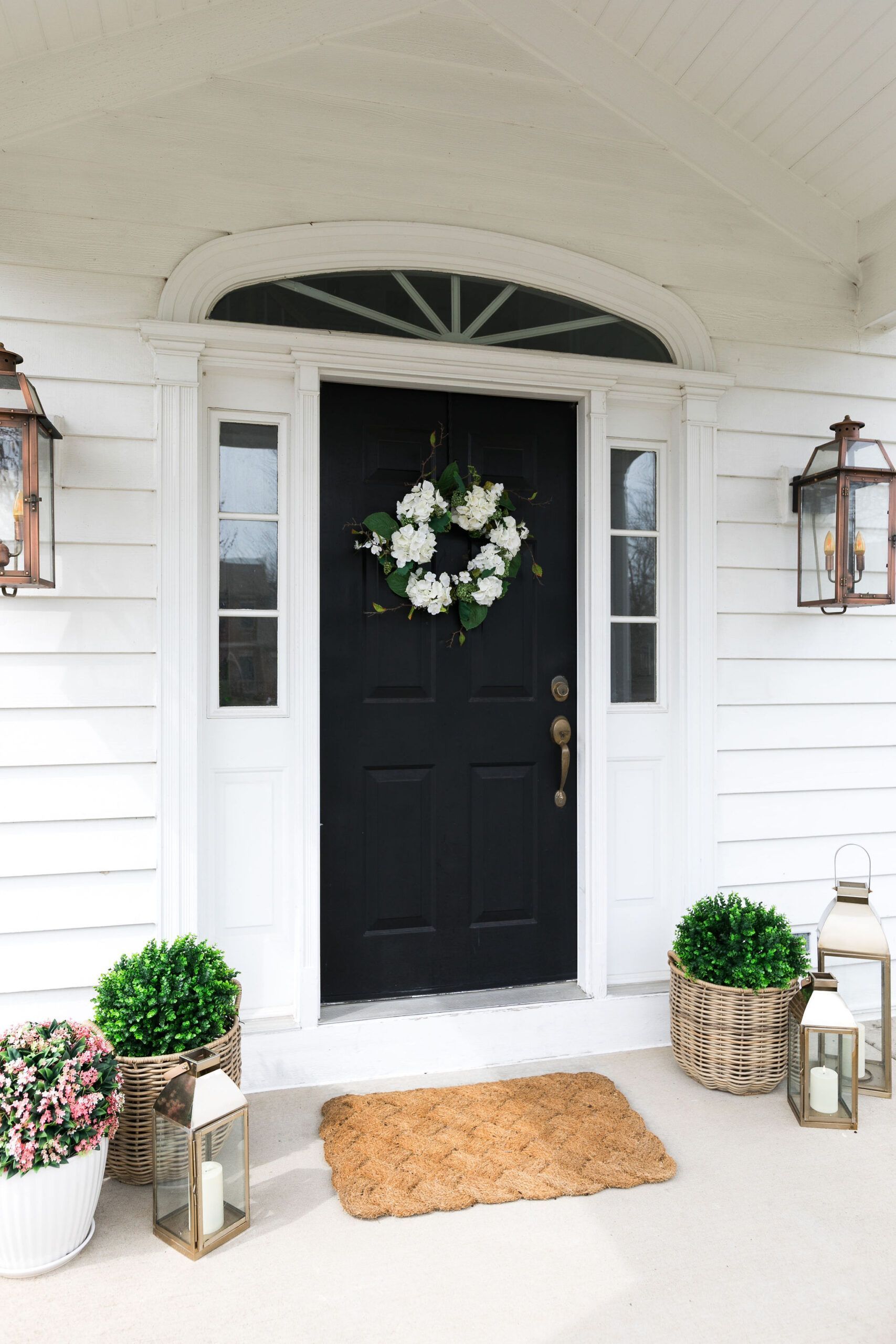 Black front door with a floral wreath on a white porch, flanked by lanterns and potted greenery.