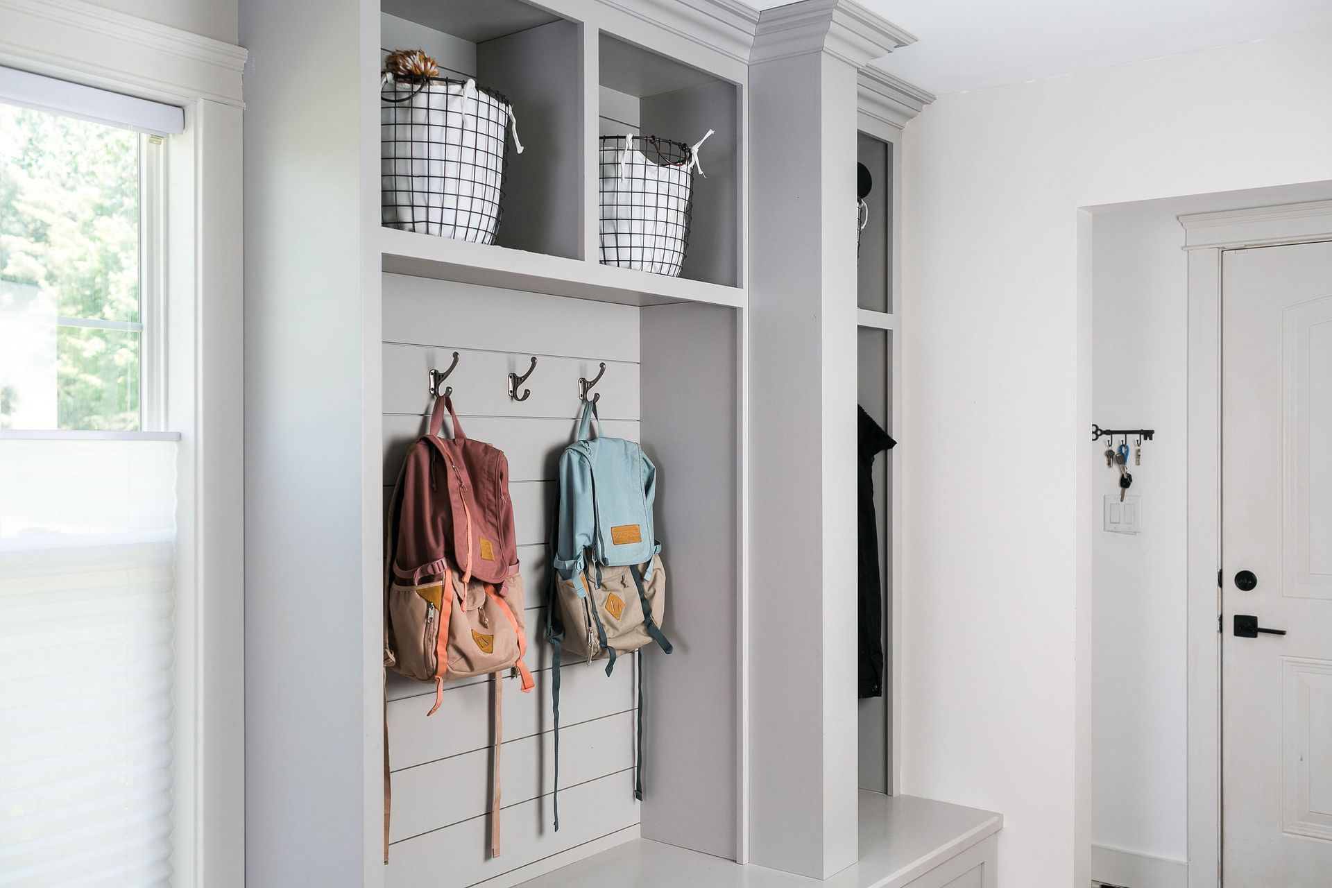 Mudroom with built-in storage, hooks holding backpacks, and baskets above. Bright and airy space, painted white and gray.