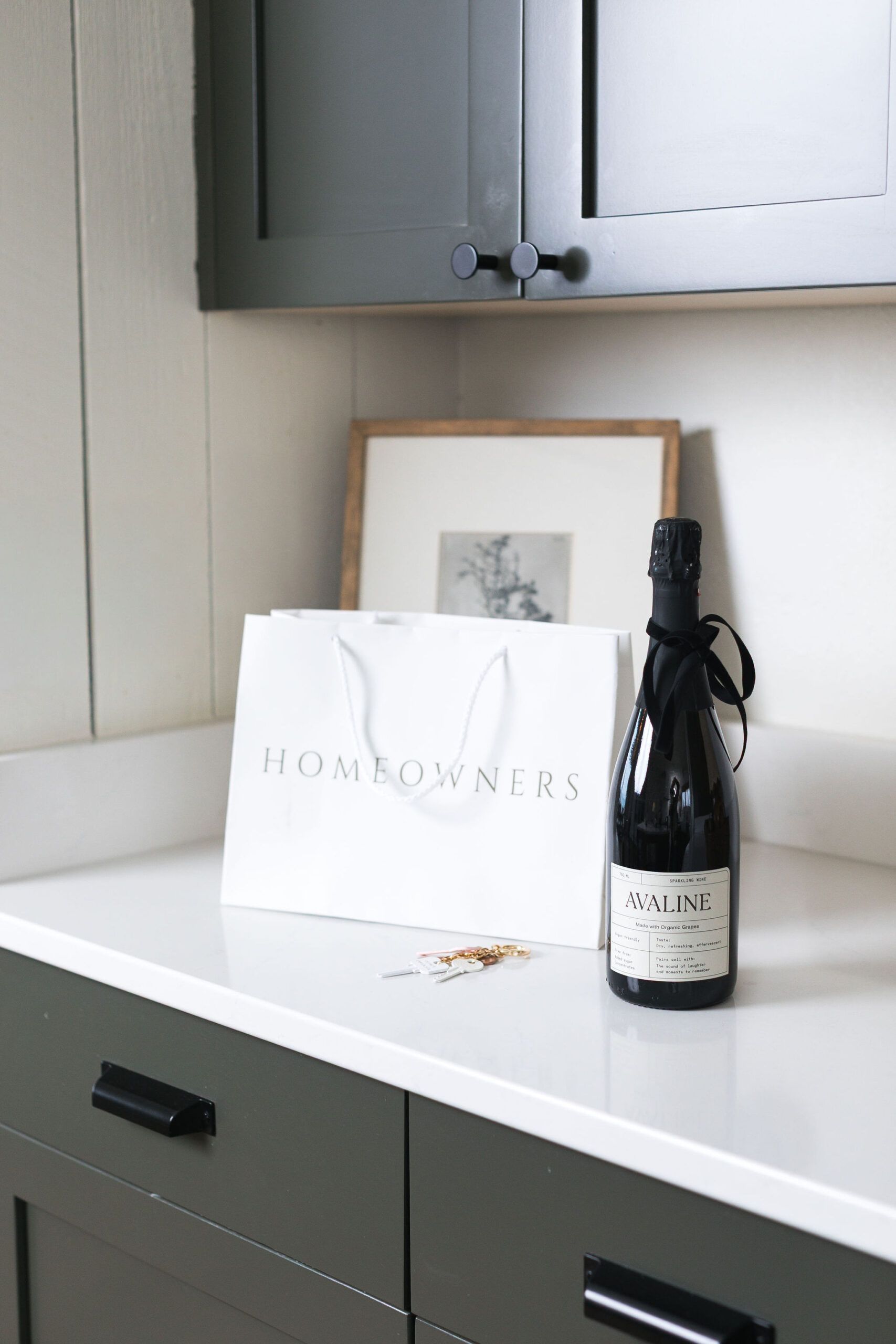 White countertop with a framed print, bag, and dark bottle beneath gray cabinets