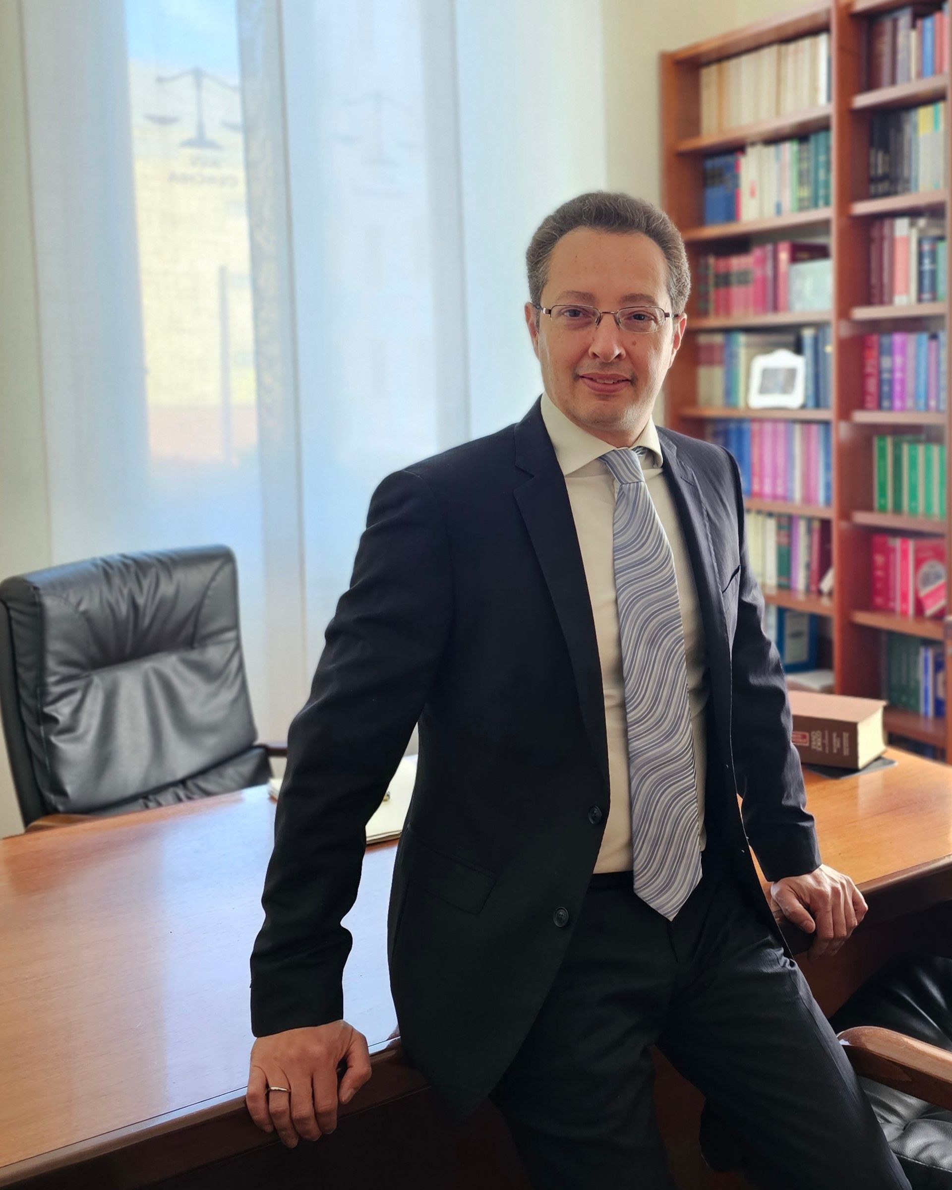 Un uomo in giacca e cravatta si appoggia a una scrivania in un ufficio con scaffali per libri.