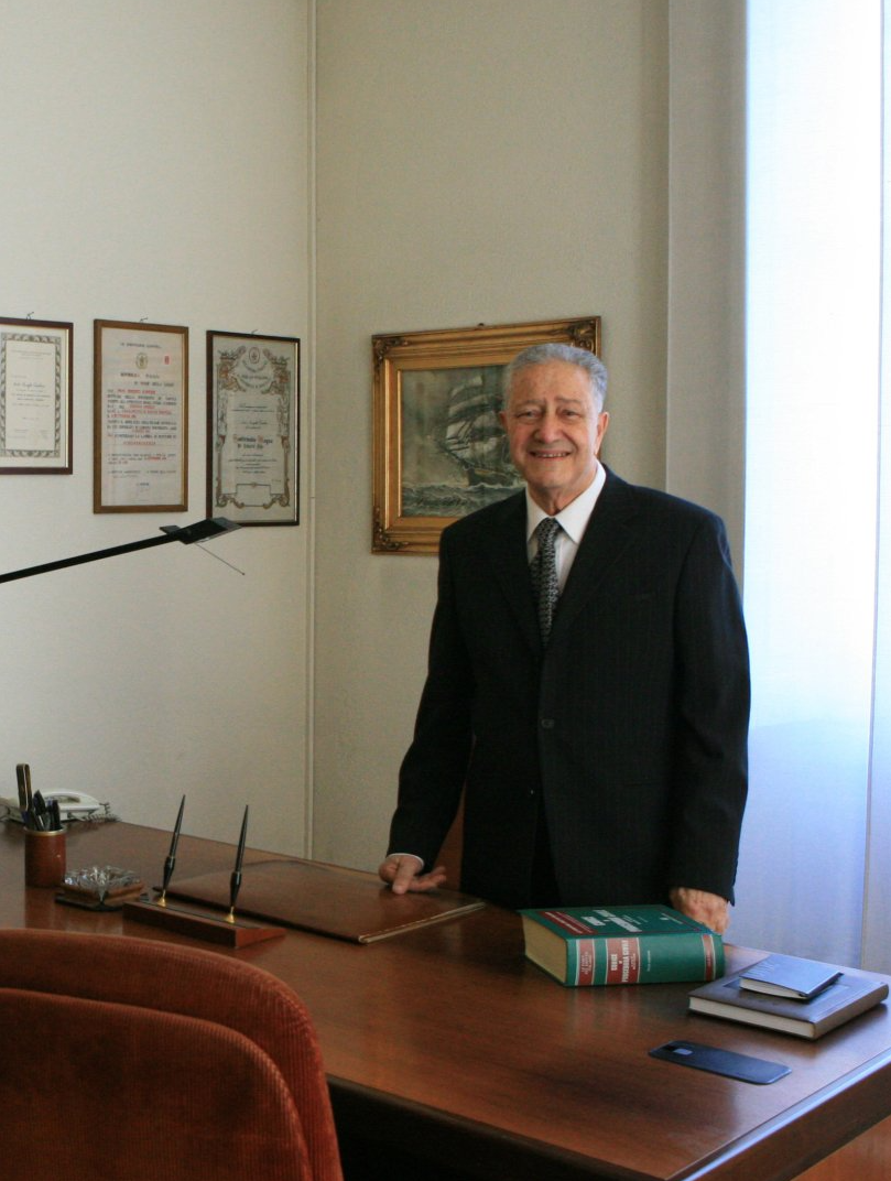 Un uomo in giacca e cravatta è in piedi dietro una scrivania in un ufficio. Dietro di lui ci sono oggetti incorniciati e un dipinto.