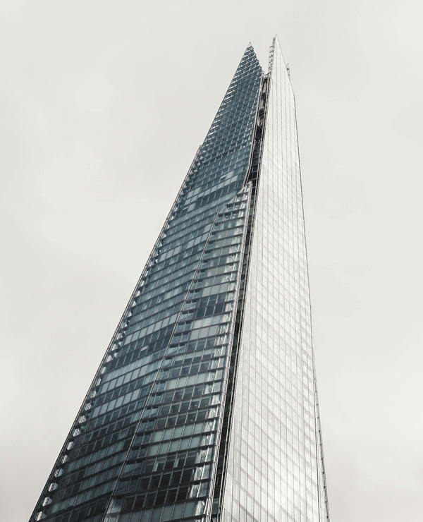 A low-angle view of the glass-clad Shard skyscraper in London, tapering to a point against a pale, overcast sky.