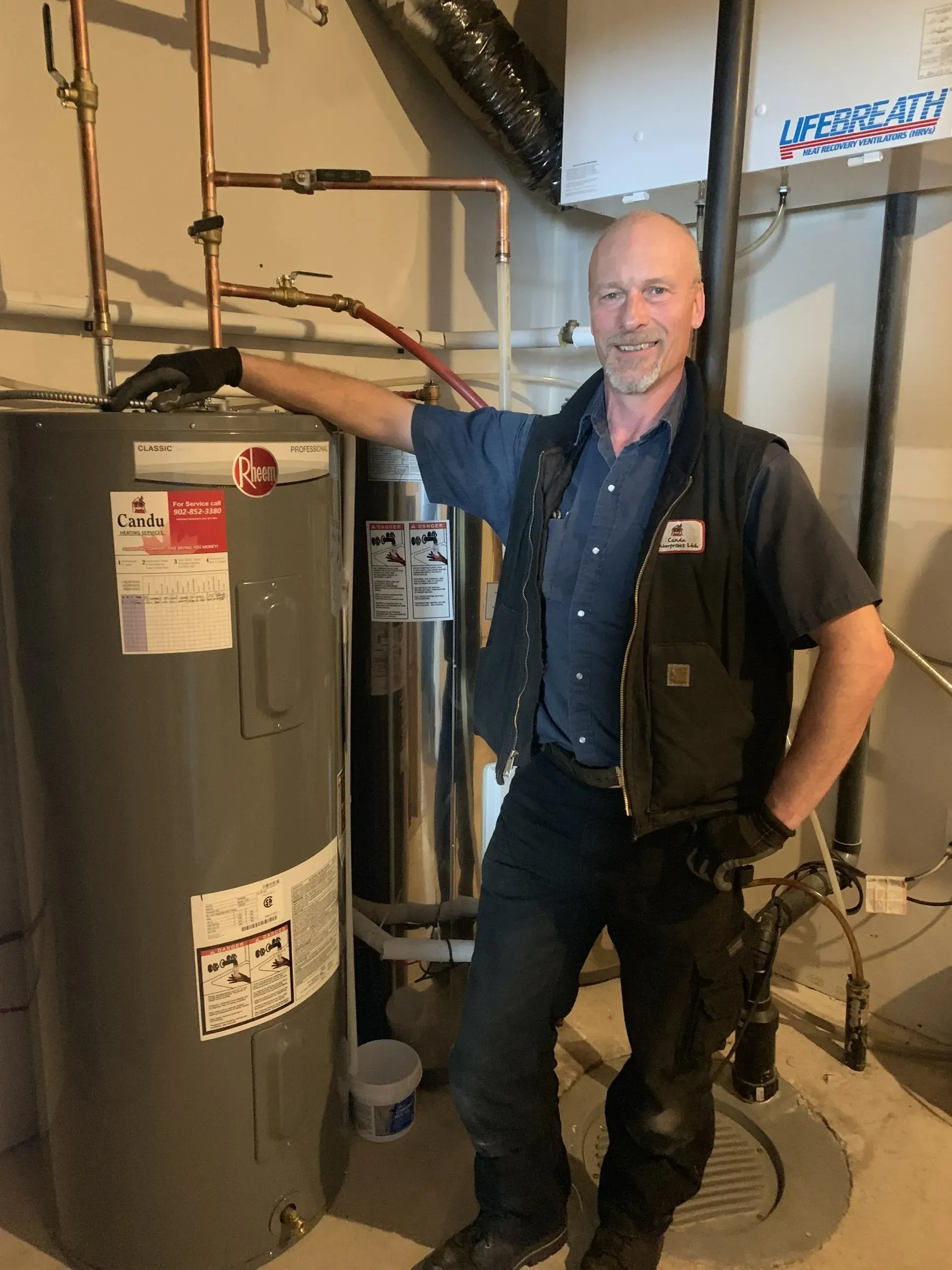 Plumber in a workshop, standing near a water heater. He wears a vest, gloves, and smiles at the camera.
