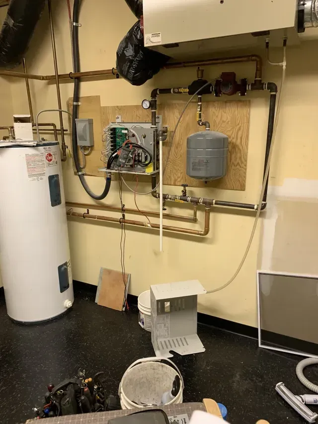 A room with a water heater and a bucket on the floor.