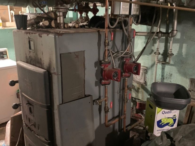 A large boiler is sitting in a basement next to a trash can.