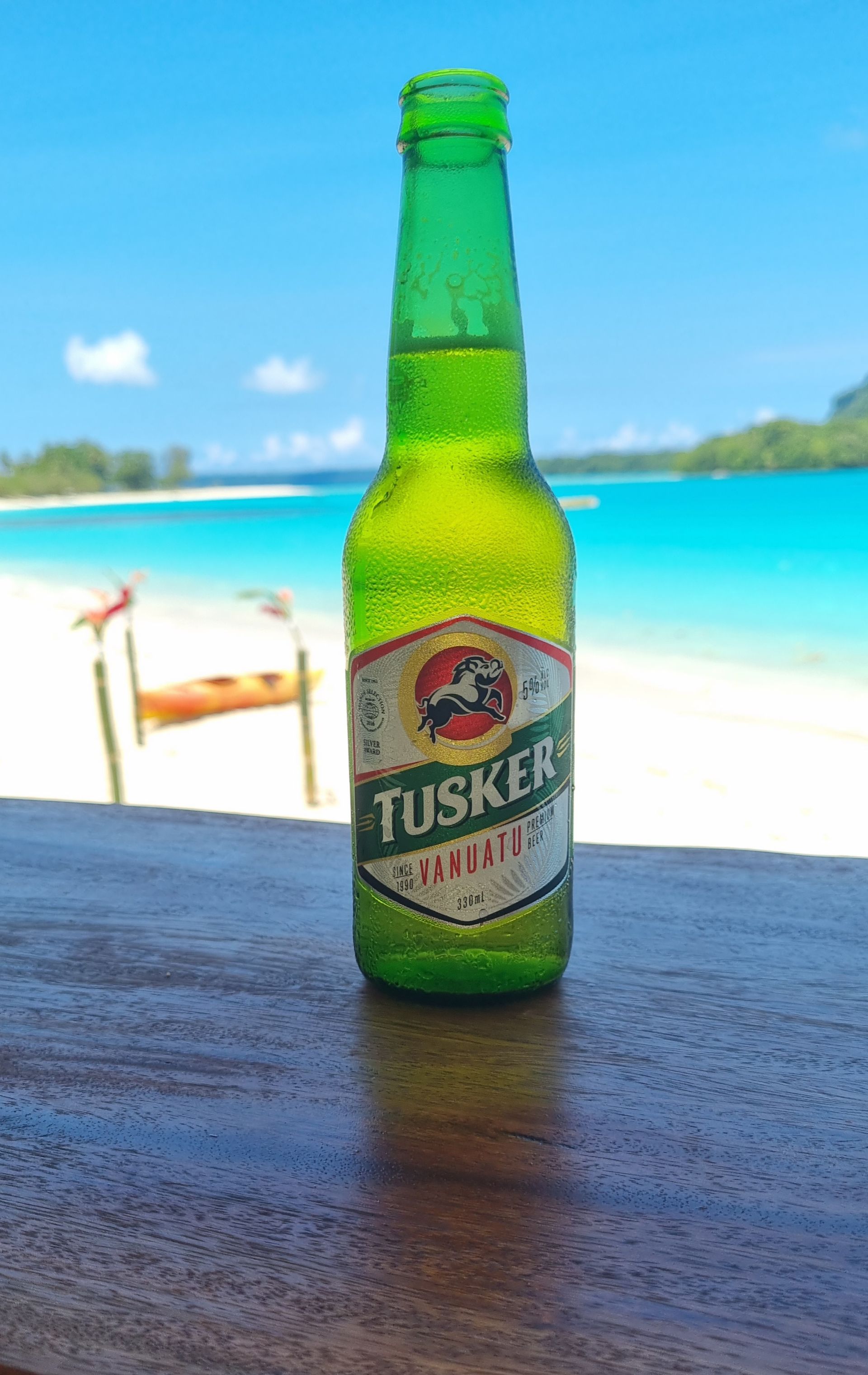 A bottle of tusker beer is sitting on a wooden table in front of a beach.